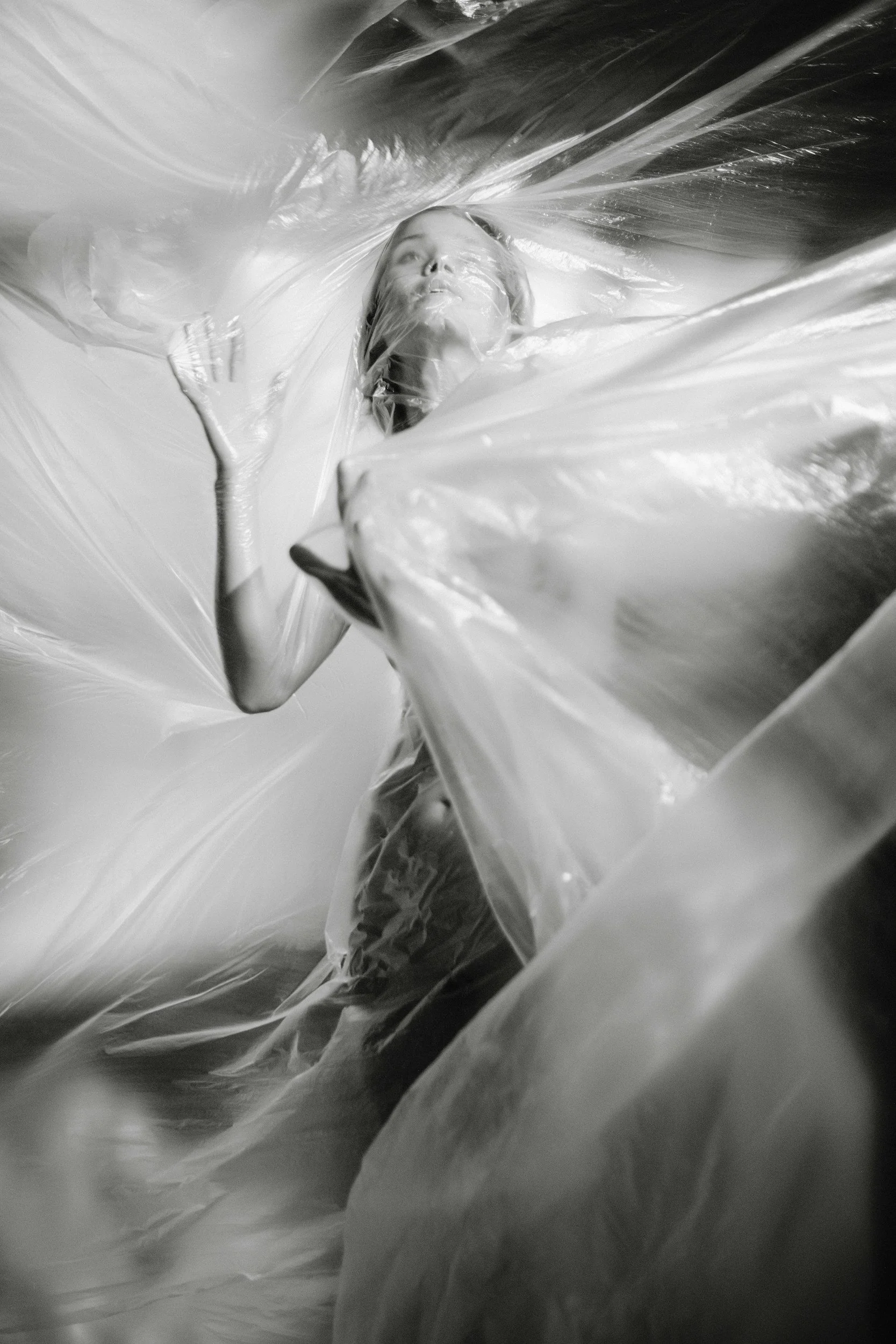 Black and white photo of a woman covered in crinkled, translucent plastic material, looking upward with her hand near her face.