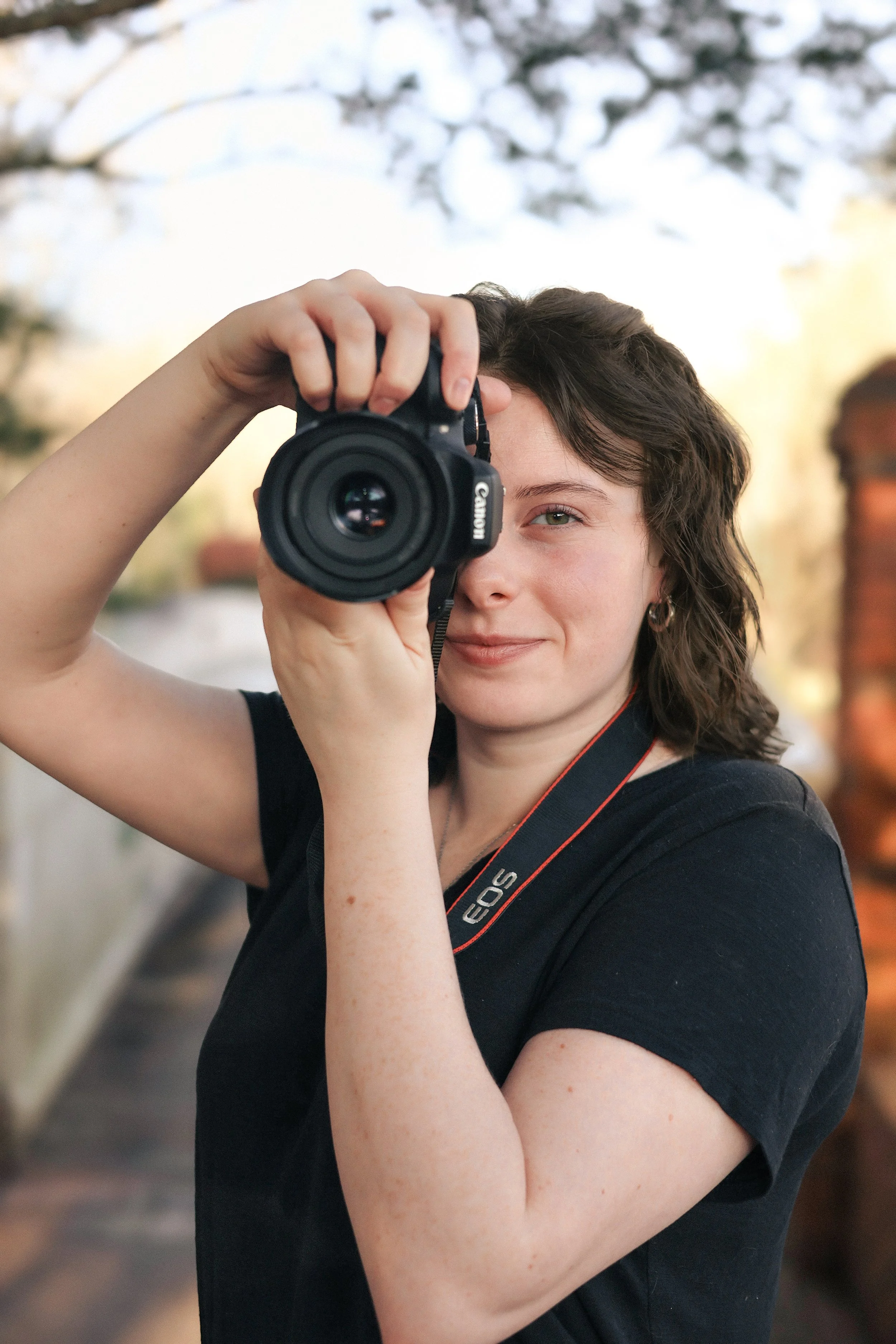 Birth photographer and overnight Doula, Katie Ashley, with curly brown hair taking a photograph with a Canon camera outdoors, with trees and a brick wall in the background.