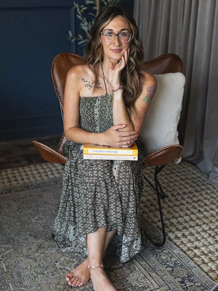 Doula Kayln Fuller sitting in a brown chair with cream pillow, wearing glasses and a printed dress, holding books on her lap, with a tattoo on her arm, and an anklet on her ankle, in a room with a patterned rug and curtains.