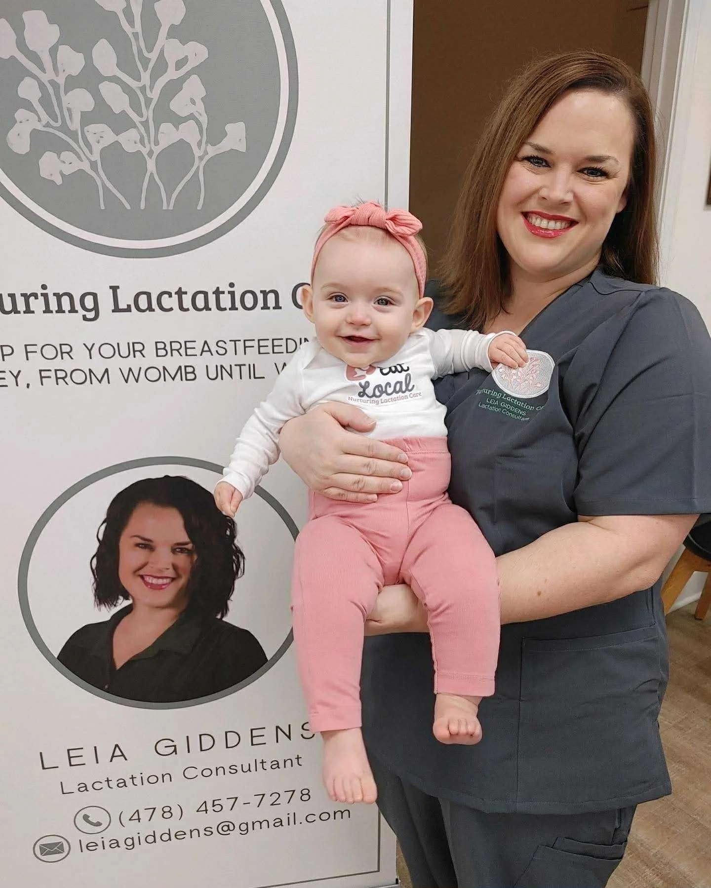 IBCLC Leia Giddens in a dark gray uniform holding a smiling baby girl in front of a sign that reads 'Nurturing Lactation' and features a portrait of a woman with dark hair and contact information.