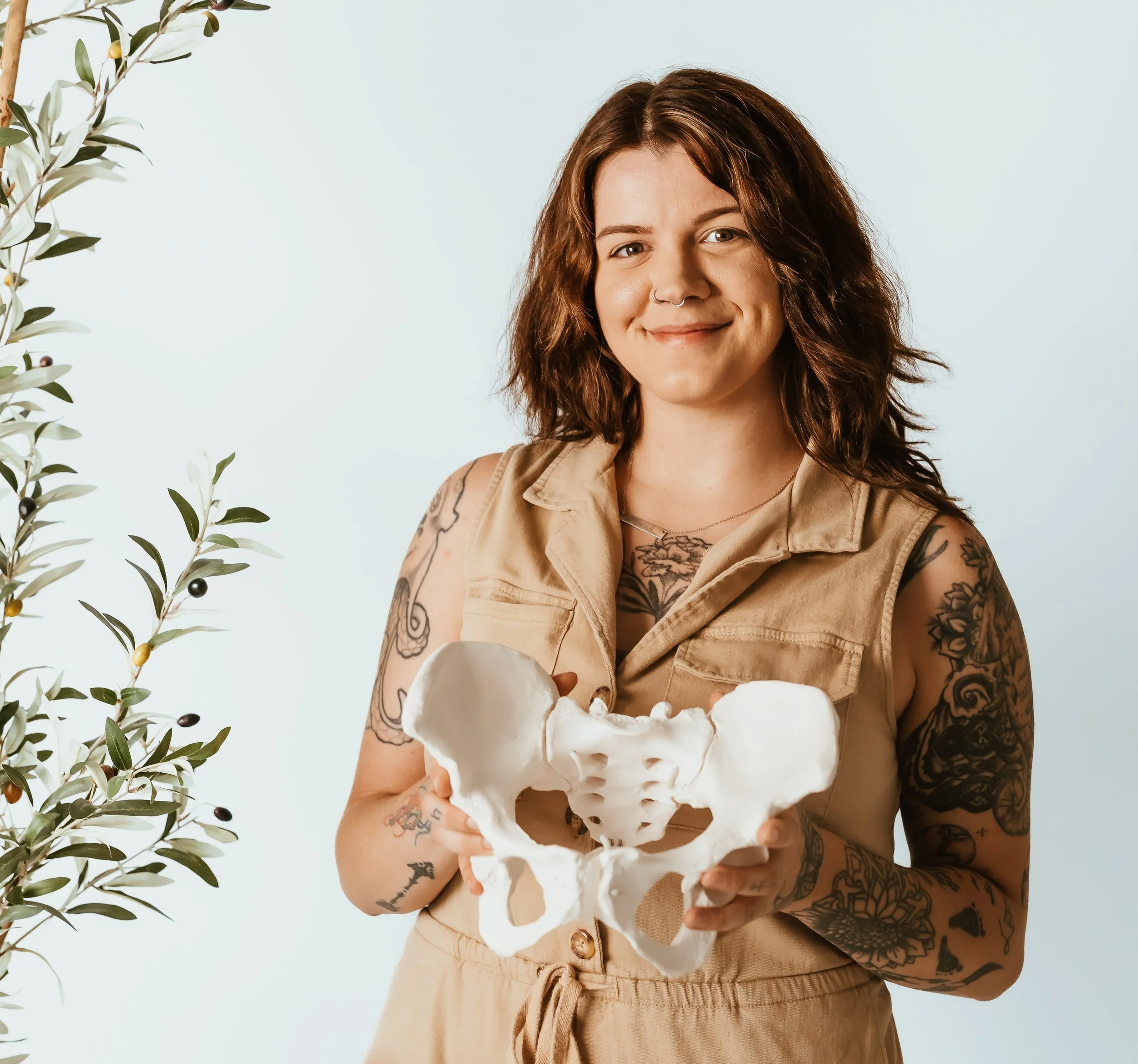 A Doula with tattoos and a septum piercing holding a model of a human pelvis against a plain light background.