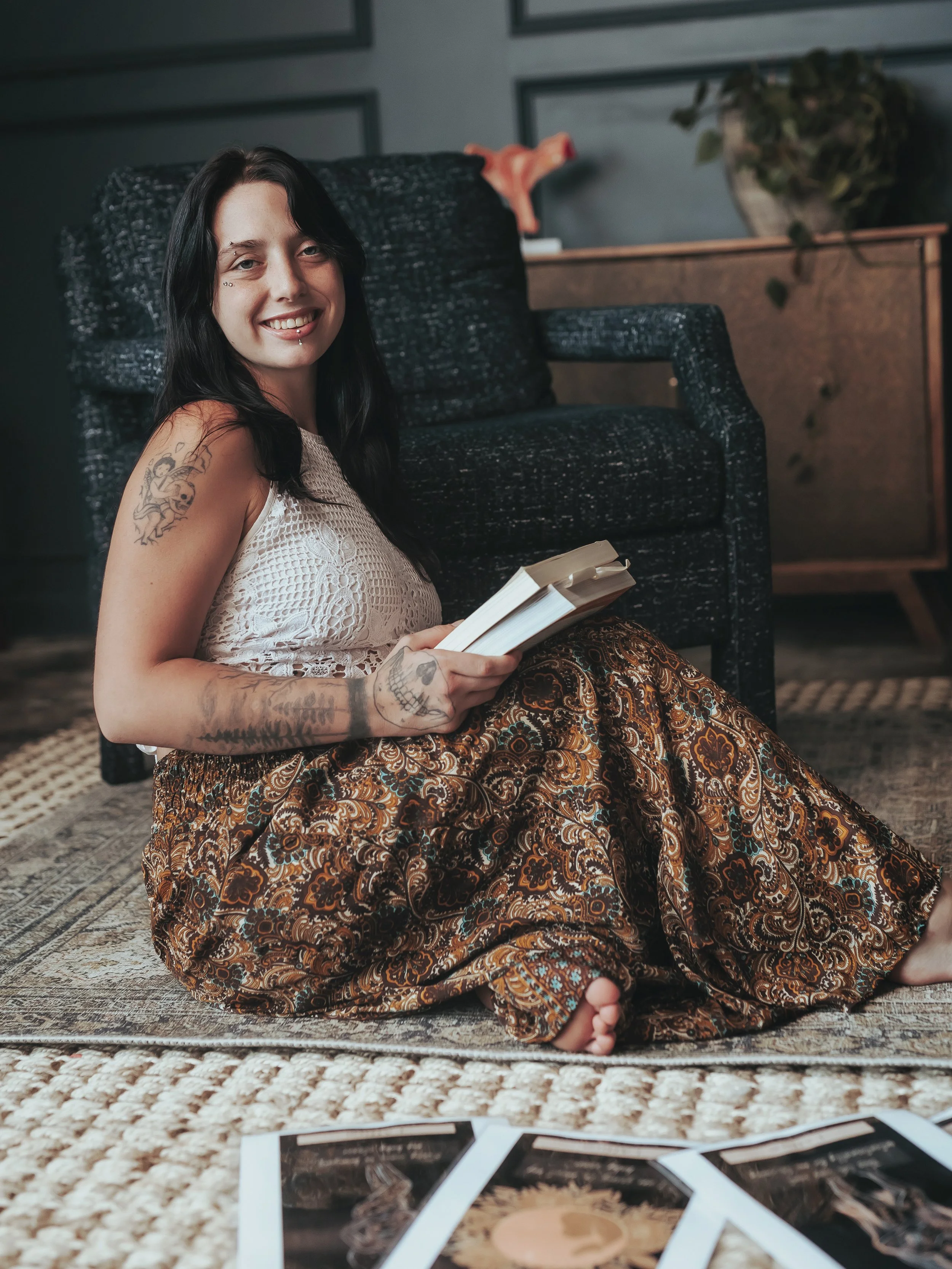Doula Enna Laflamme with tattoos, dark hair, and piercings sitting on a rug, smiling, holding an open book, with birth affirmations on the floor around her,