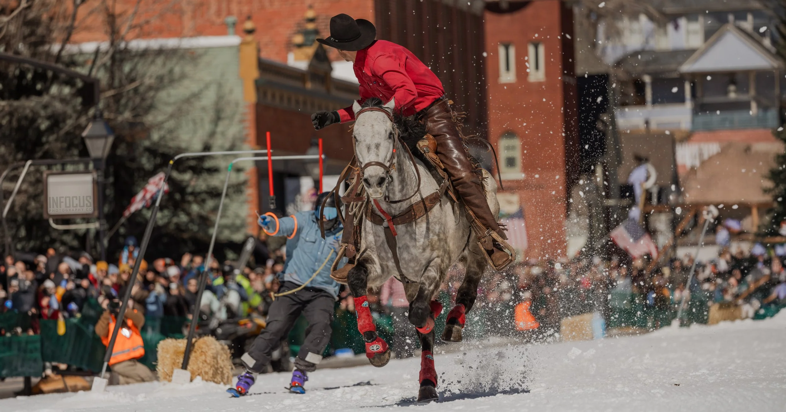 Skijoring competitor in a red jacket rides a horse through the snow as spectators watch.
