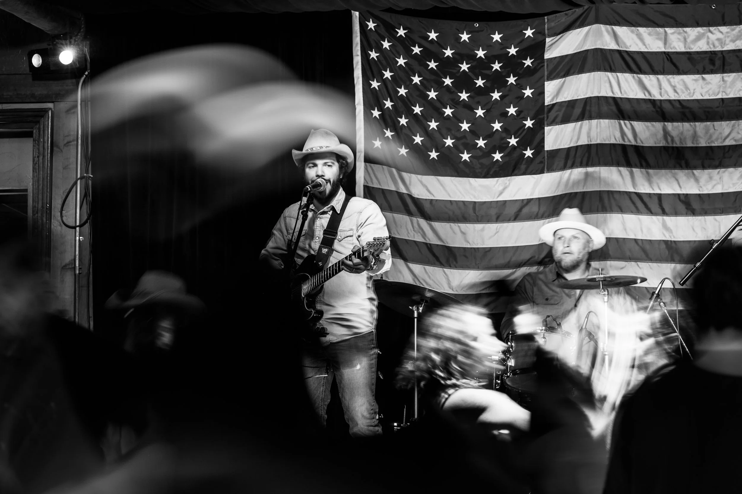 Musician sings into a microphone with an acoustic guitar in a black-and-white stage portrait.