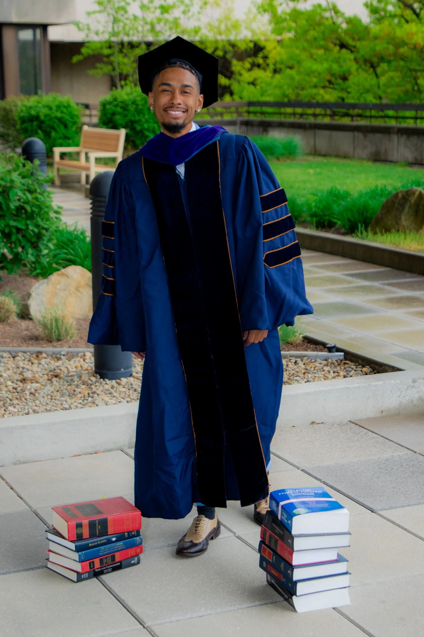 A young man in a graduation cap and gown stands outdoors on a paved walkway, smiling. There are two stacks of books on the ground beside him, one on each side.
