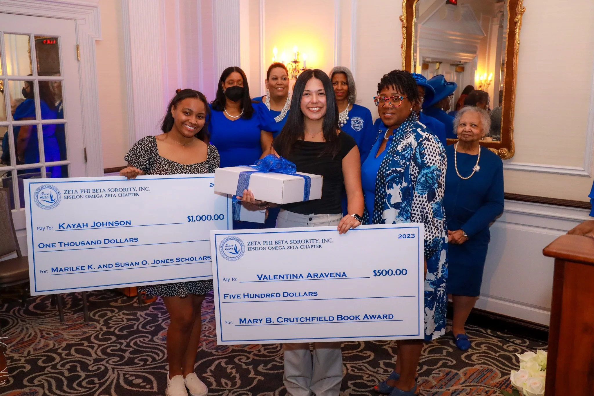 Group of women at an awards ceremony, holding oversized checks and a gift box, with elegant decor including a large mirror and chandelier in the background.