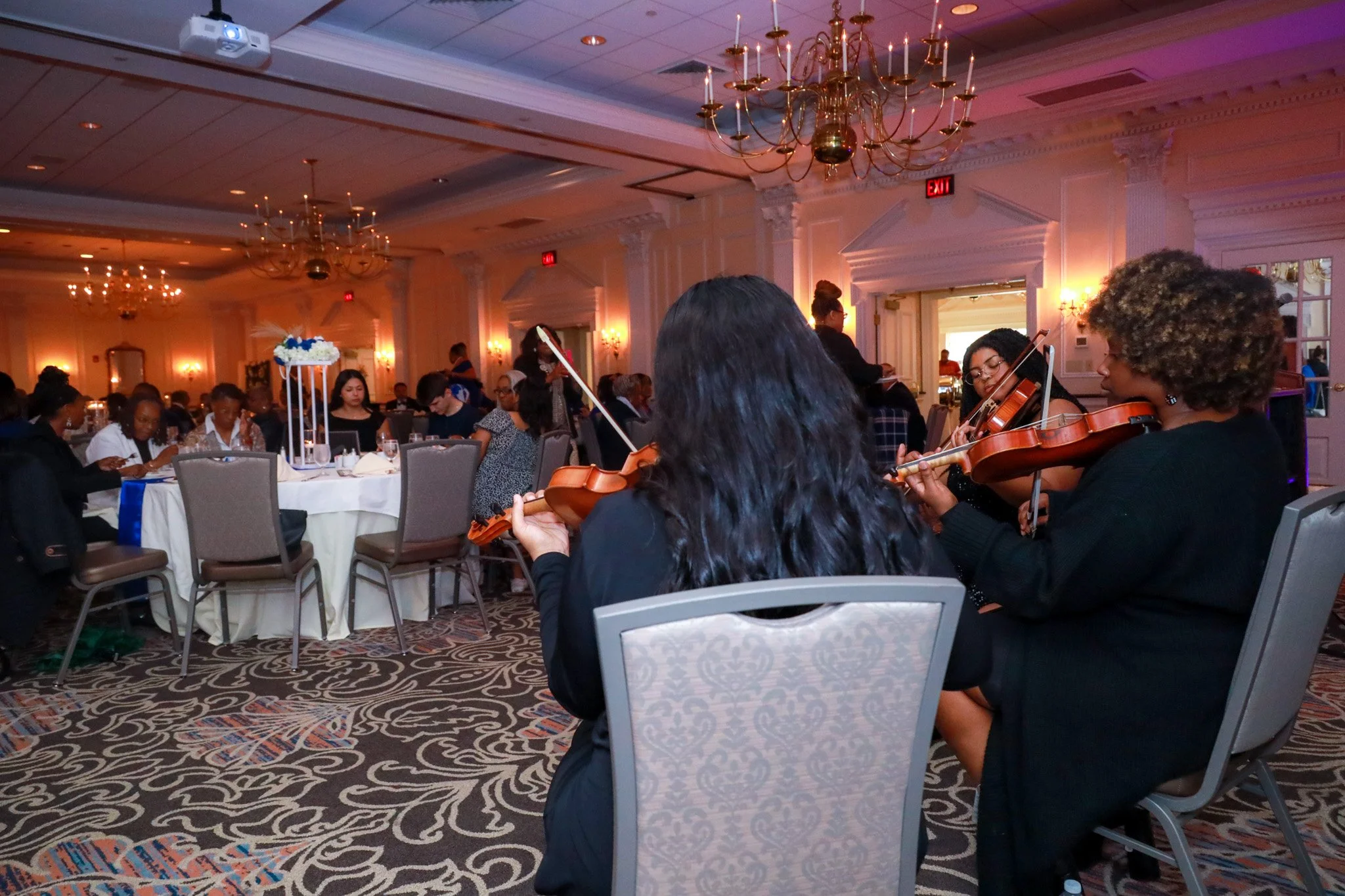 A group of female musicians playing violins at an indoor event with elegant chandeliers and well-lit decor, while guests sit at round tables in the background.