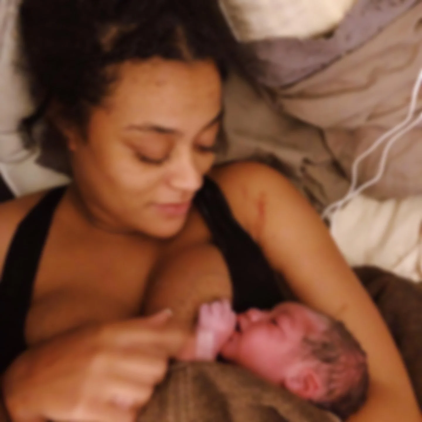 A woman with curly hair lying in bed, smiling, holding a baby close to her chest, with visible scratches on her arm.
