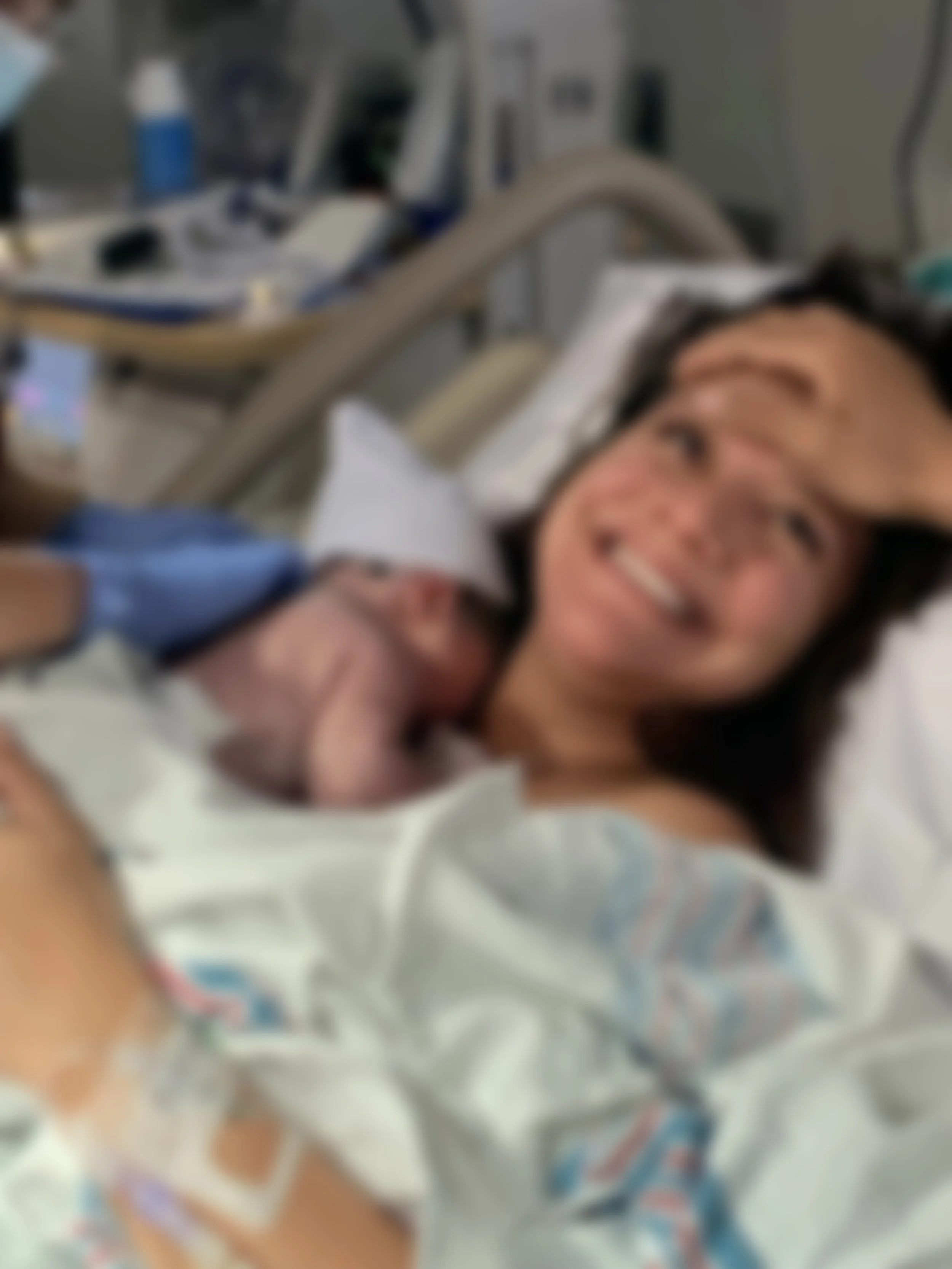 A woman lying in a hospital bed holding a newborn baby, both smiling, with medical equipment and blankets in the background.