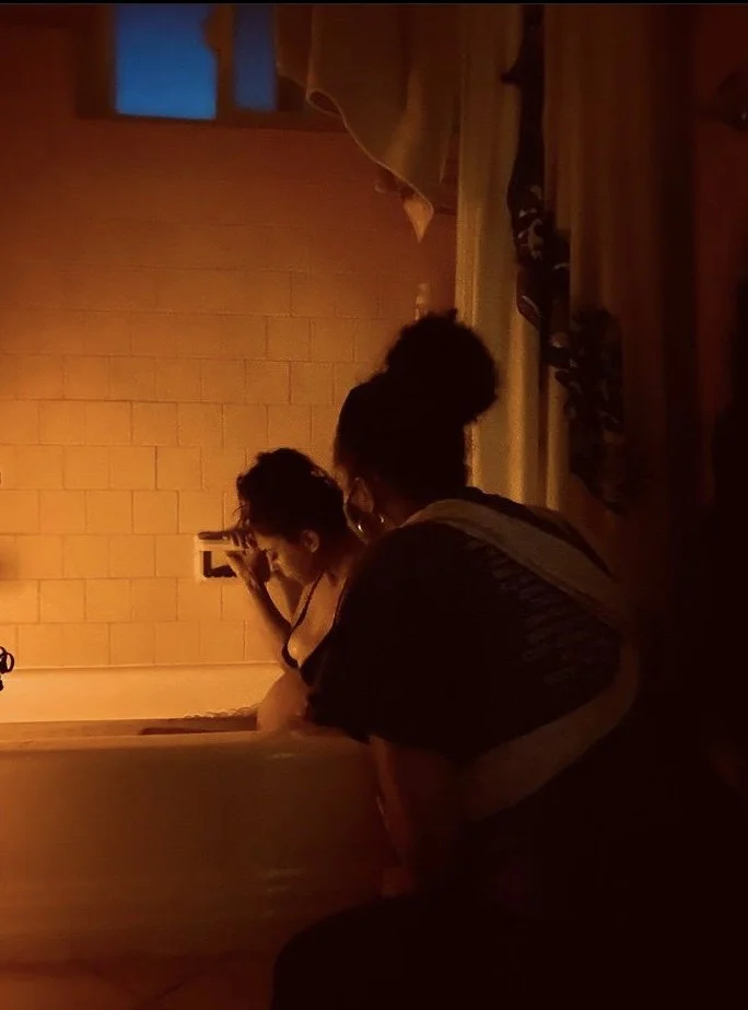 Two people sitting in a dimly lit room, one adjusting a light switch or outlet on a brick wall, the other facing away from the camera, with a small window showing a blue sky in the background.