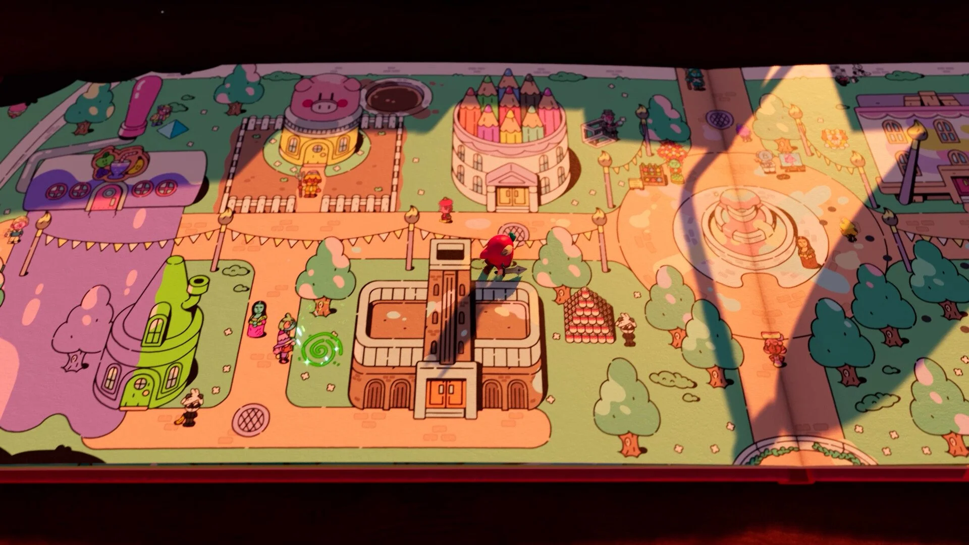 A colorful, top-down view of a storybook level from The Plucky Squire. The character Jot stands in a vibrant town square with pencil-shaped buildings, a fountain, and stylized trees. Dramatic shadows stretch across the page.