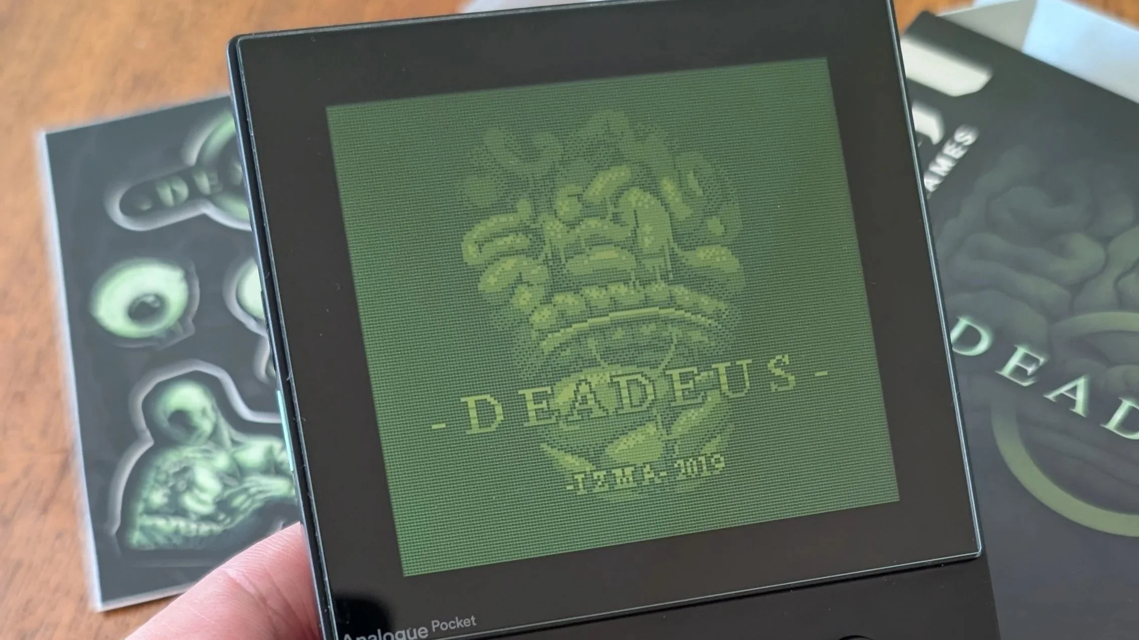 An Analogue Pocket handheld running the indie horror game Deadeus, showcasing the physical cartridge and the GB Studio pixel art aesthetic reviewed by Low Five Gaming.