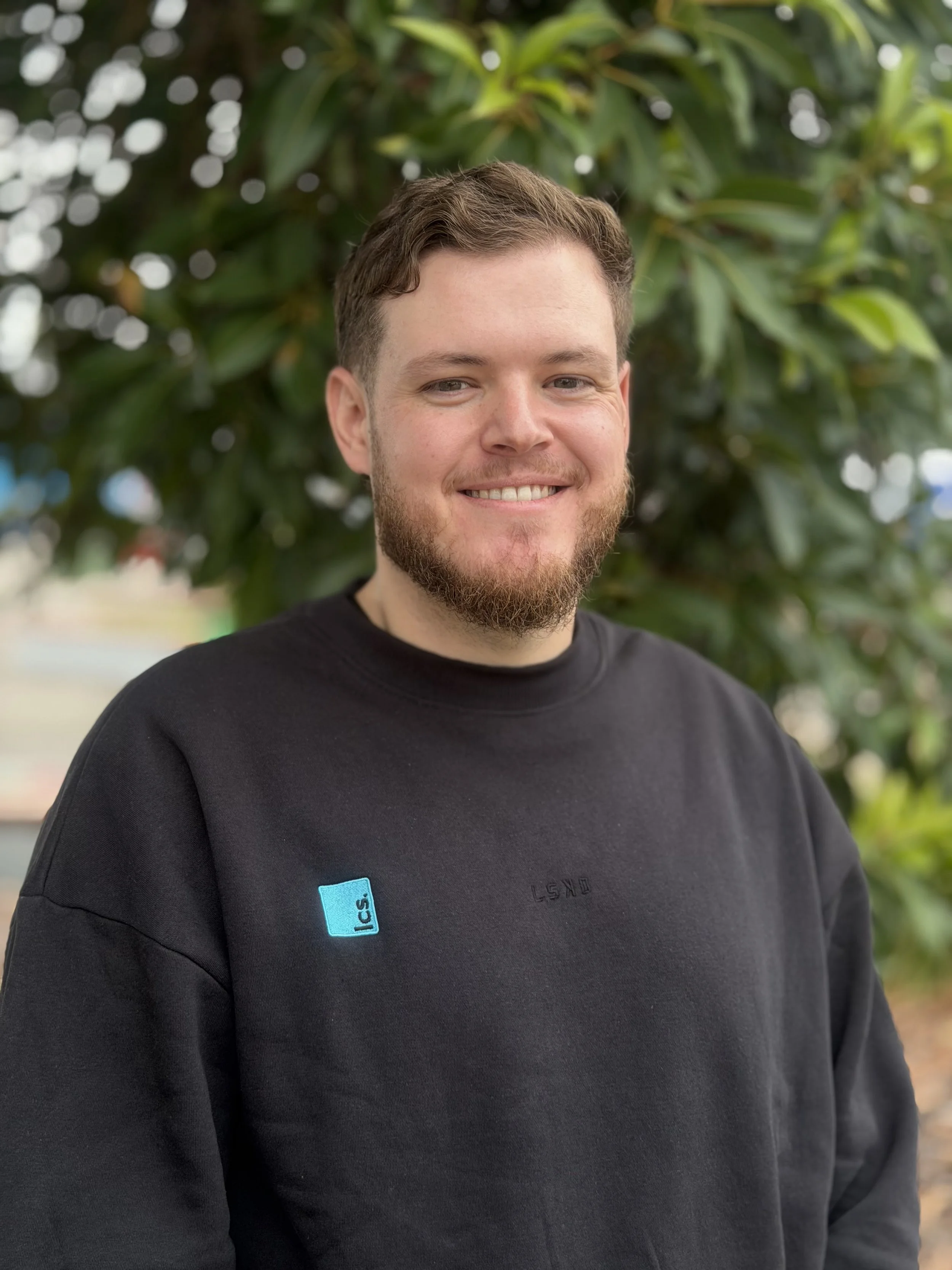 A bearded man is standing facing the camera, smiling. He is wearing a black round-neck jumper. There is a green leafy tree in the background.