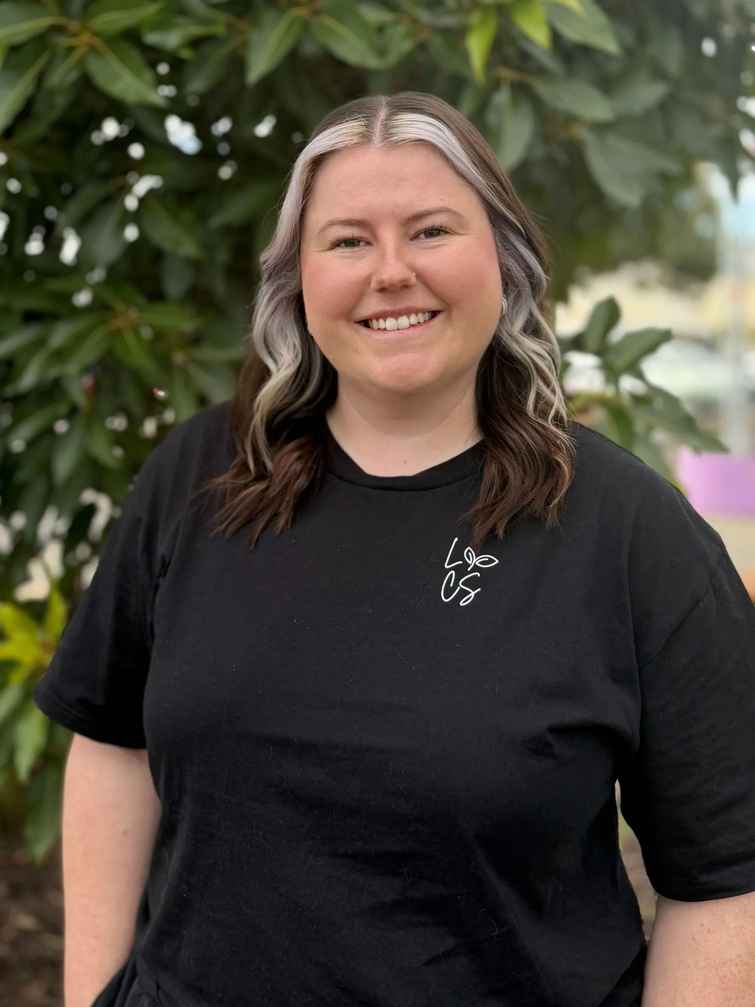 A woman with shoulder-length dark brown hair, smiling, wearing a black polo shirt with a small blue logo, standing outdoors with trees and a fence in the background.