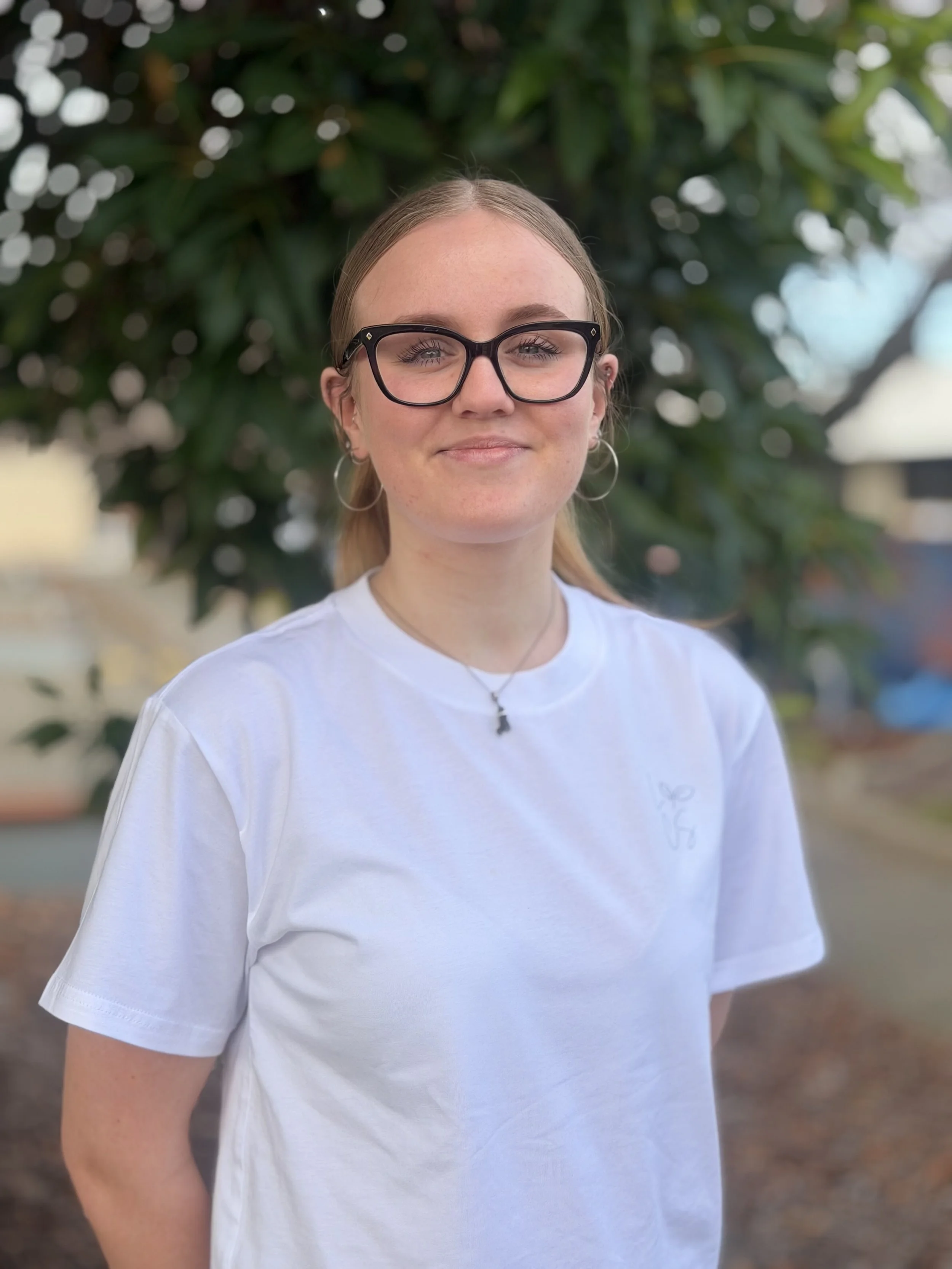 A woman standing outdoors on a sunny day, smiling, wearing a white t-shirt and dark rimmed glasses, with a tree in the background.
