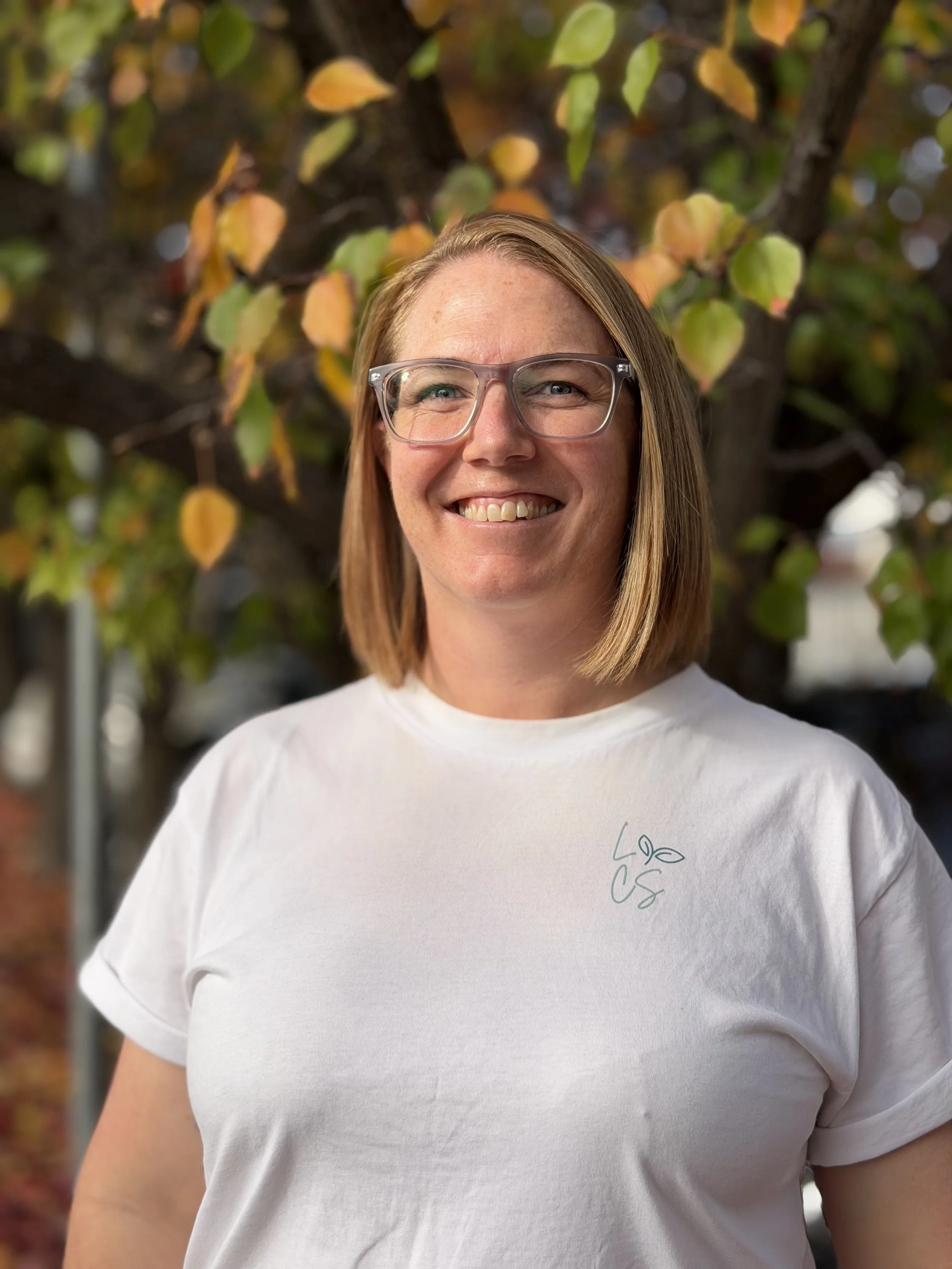 A woman with shoulder-length brown hair, wearing glasses and a black polo shirt with 'linary community services' embroidered on the sleeve, posing outdoors with crossed arms and a smile, in front of green trees and a yellow fence.