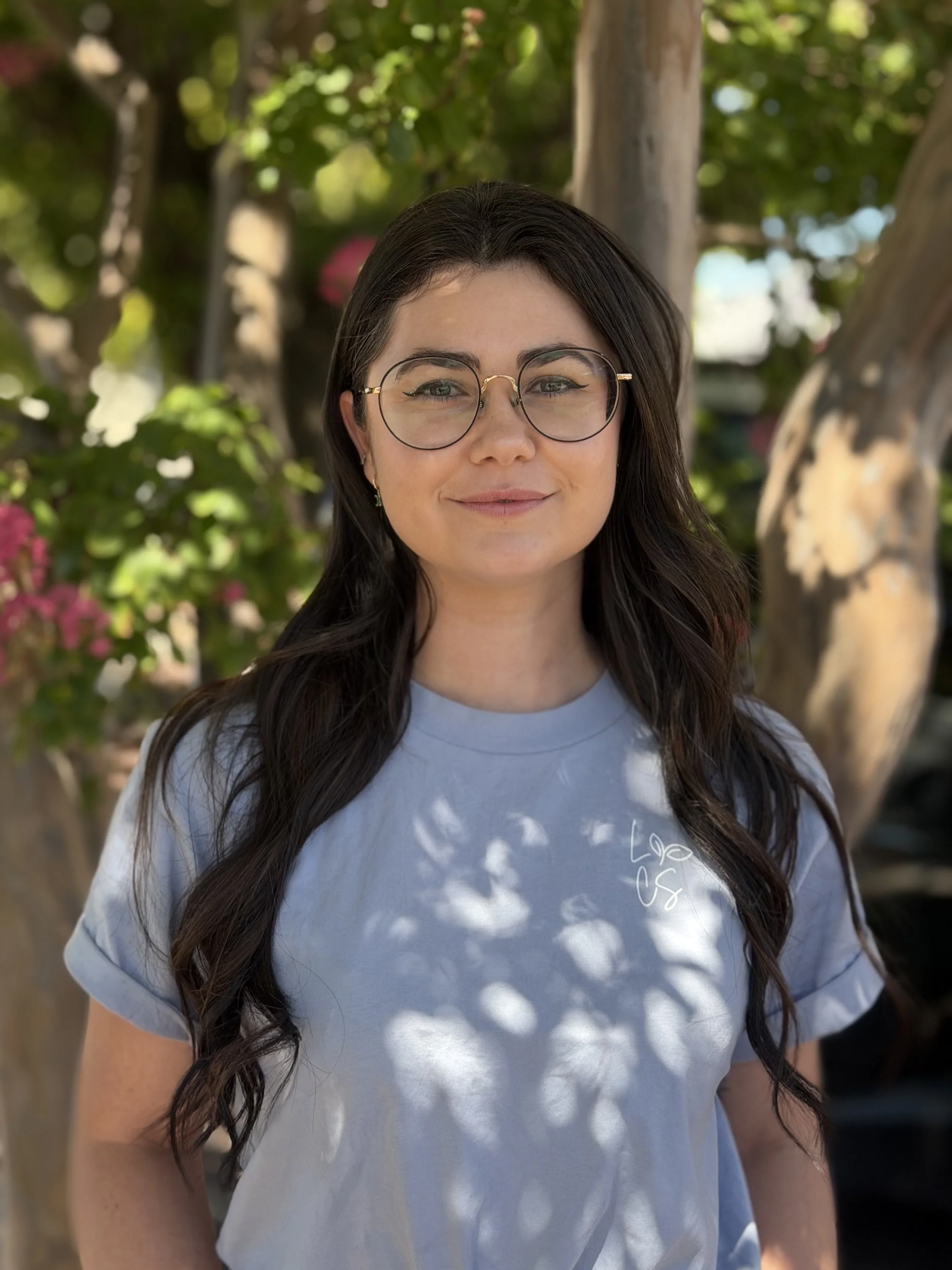 A woman with long dark hair, smiling at the camera with a green, leafy tree in the background.