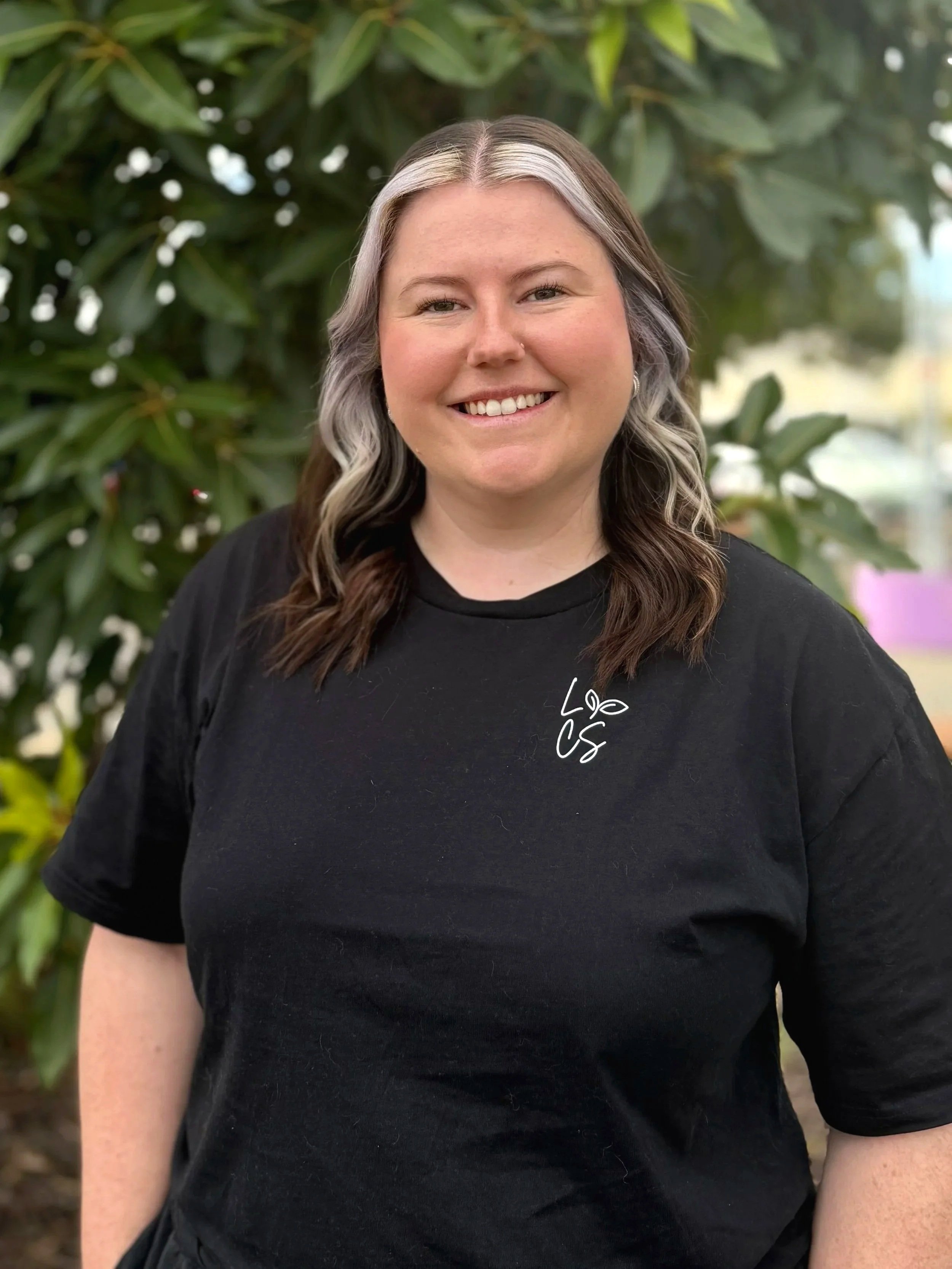 A woman with shoulder-length dark brown hair, smiling, wearing a black polo shirt with a small blue logo, standing outdoors with trees and a fence in the background.