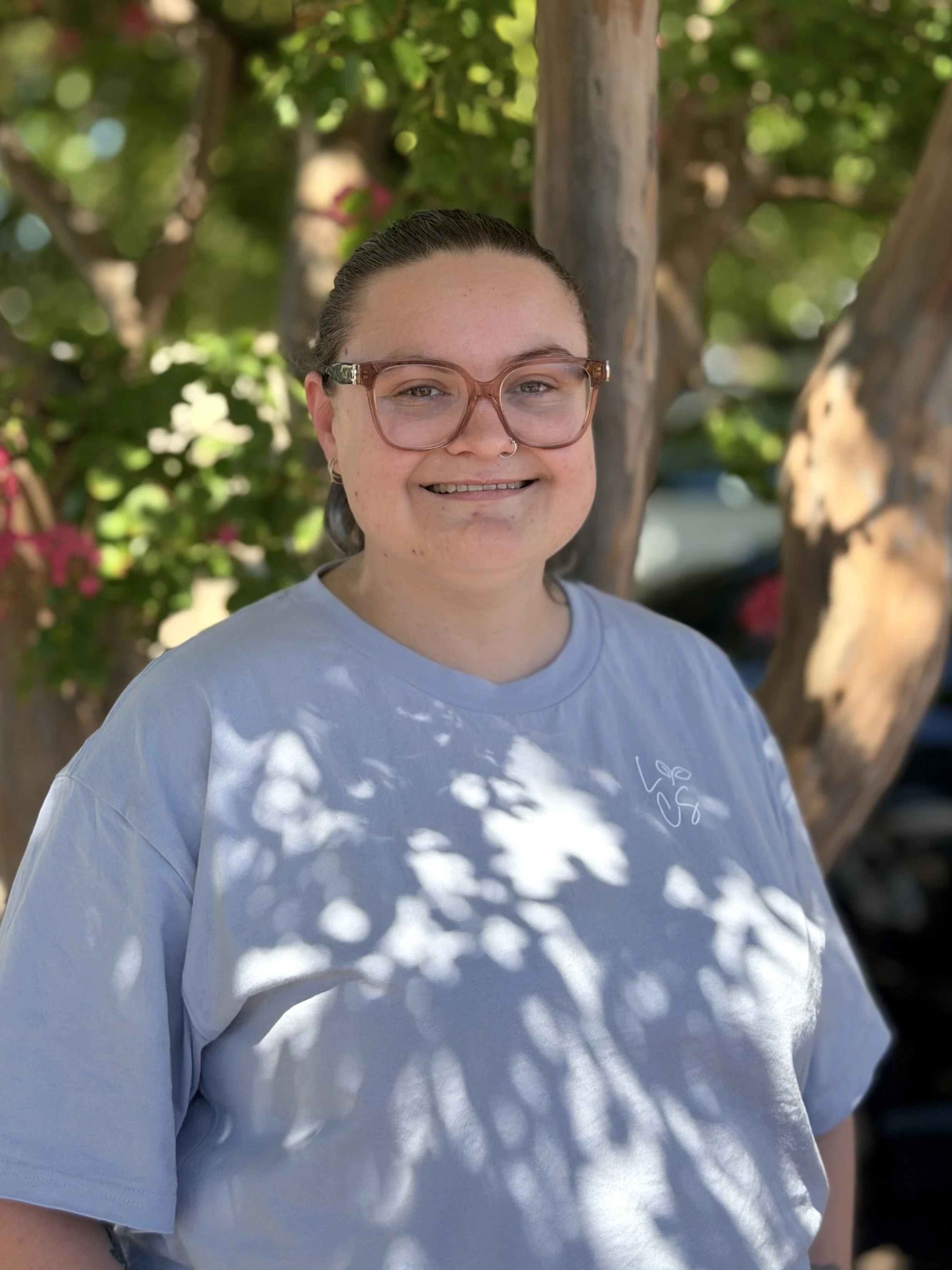A person with hair tied back, wearing glasses, smiling towards camera with green, leafy tree in the background.