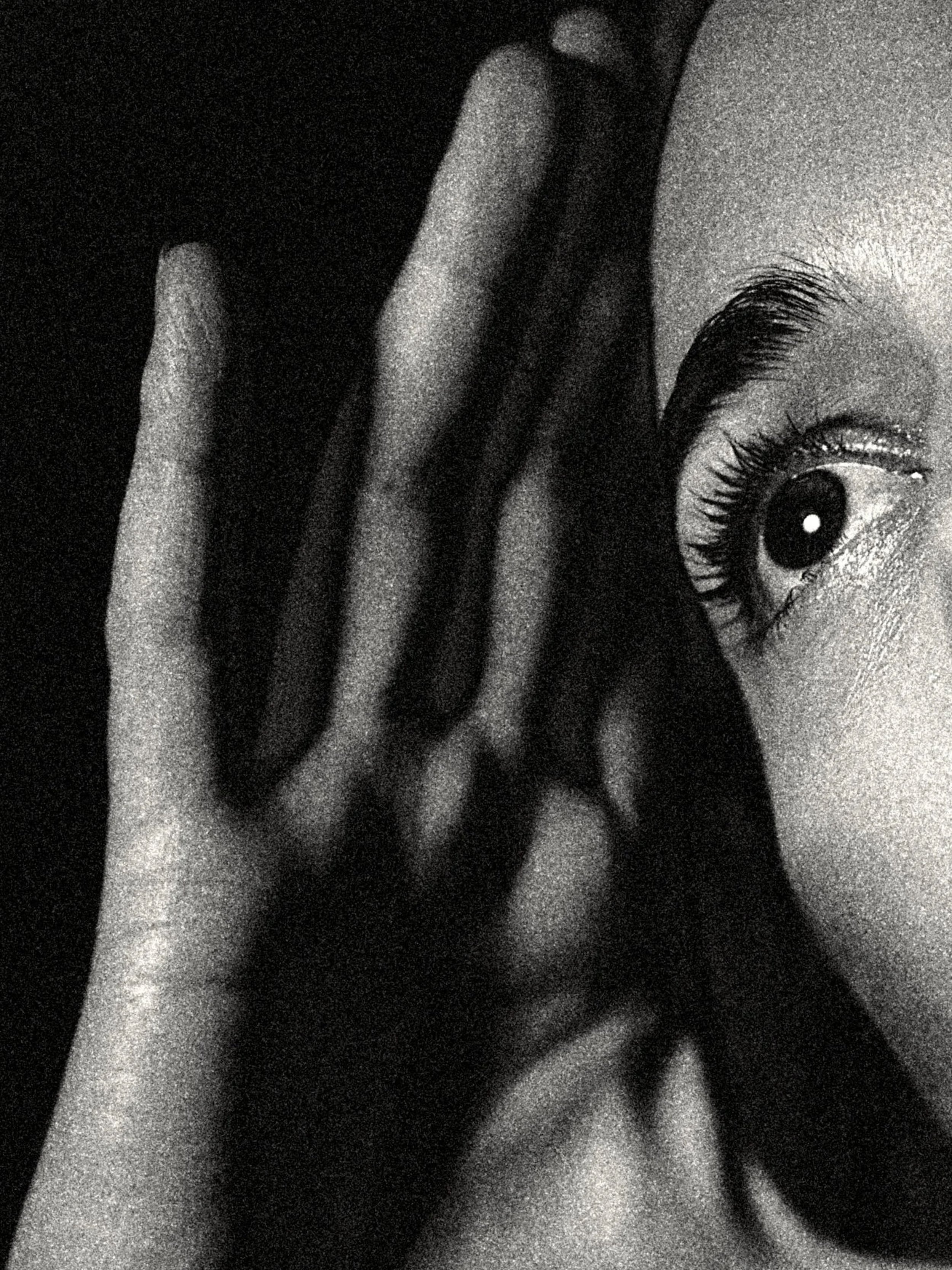 Extreme close-up black-and-white portrait emphasizing one wide-open eye and a hand lifted near the ear, suggesting listening, secrecy, and heightened attention.