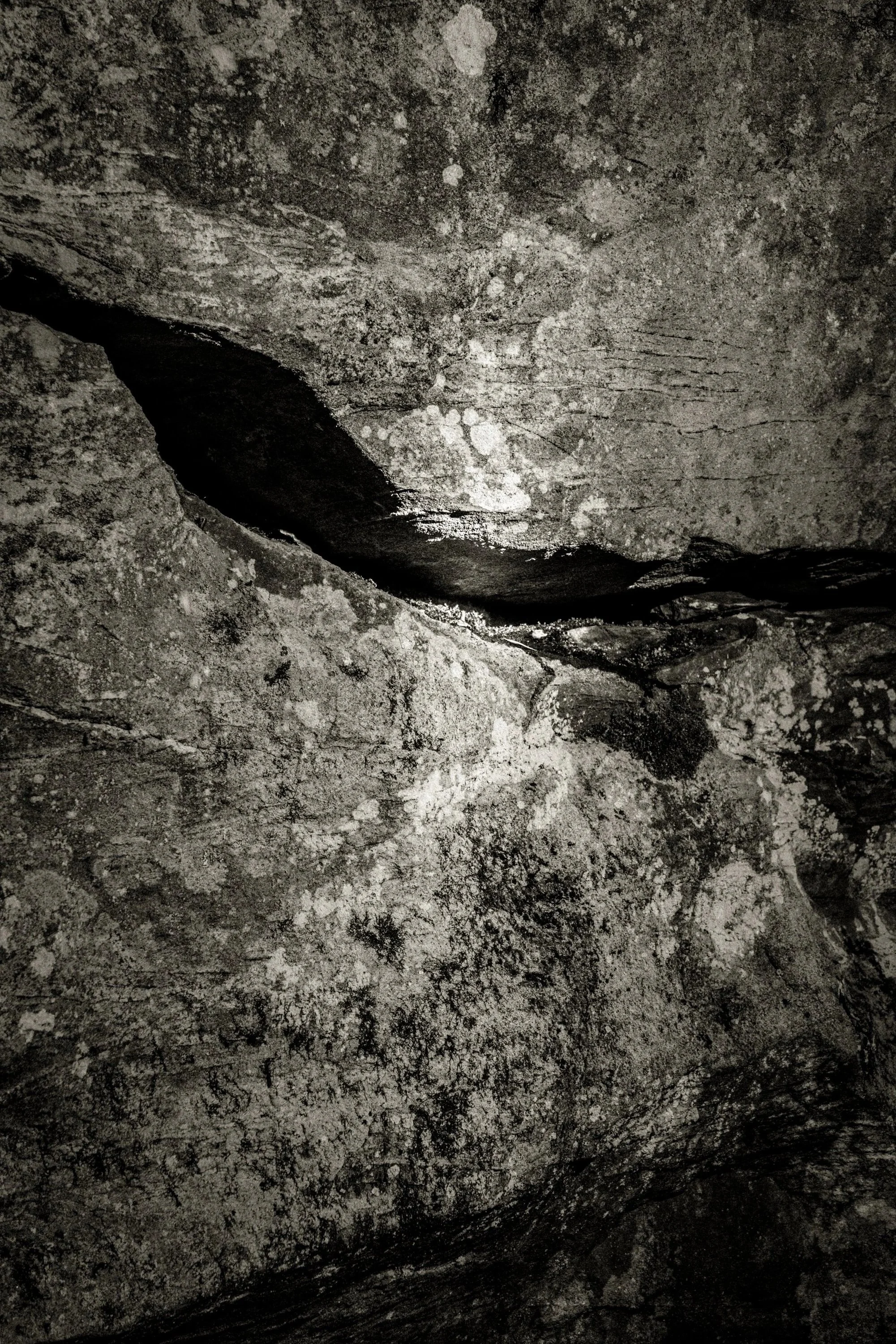 High-contrast black-and-white close-up of rock textures and a dark crevice with a faint reflective glint, emphasizing erosion, hidden depth, and geologic abstraction.