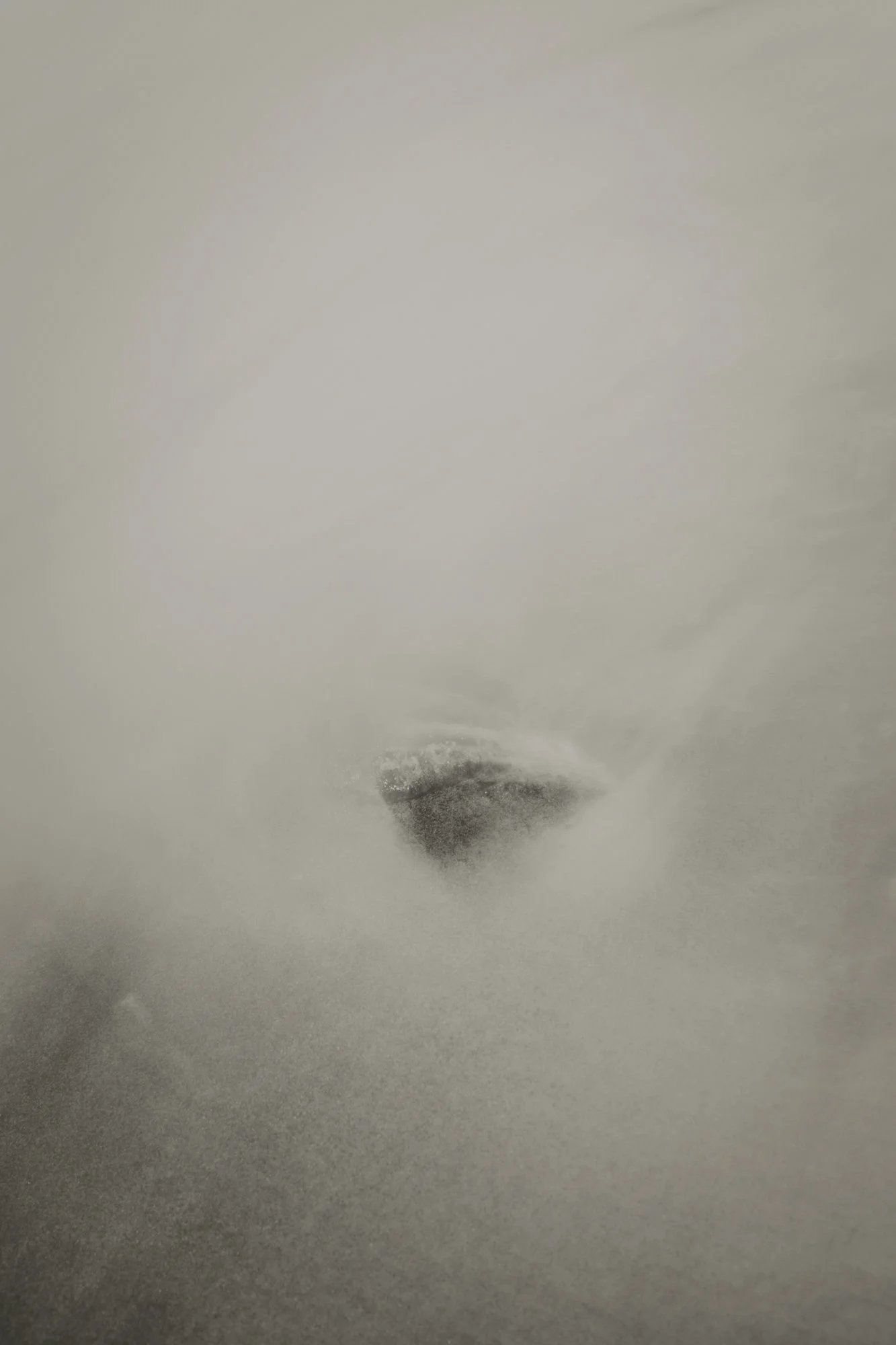 Minimalist black-and-white image of misty water and spray surrounding a small dark rock, with vast negative space suggesting silence, void, and depth.