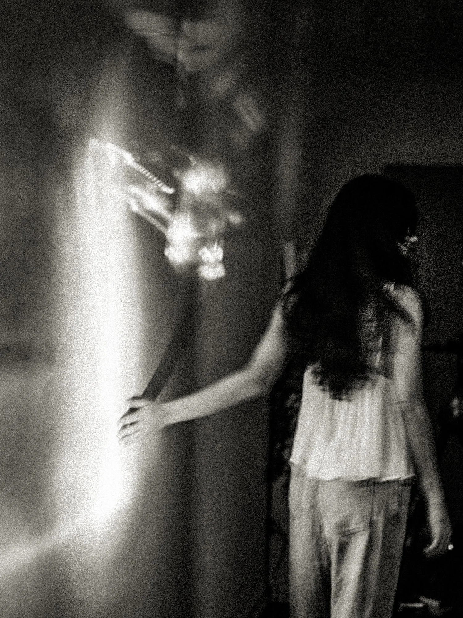 High-grain black-and-white scene of a figure reaching toward a bright vertical beam of light in a dark room, layered with ghostly reflections—an atmospheric image of threshold, revelation, and contact.