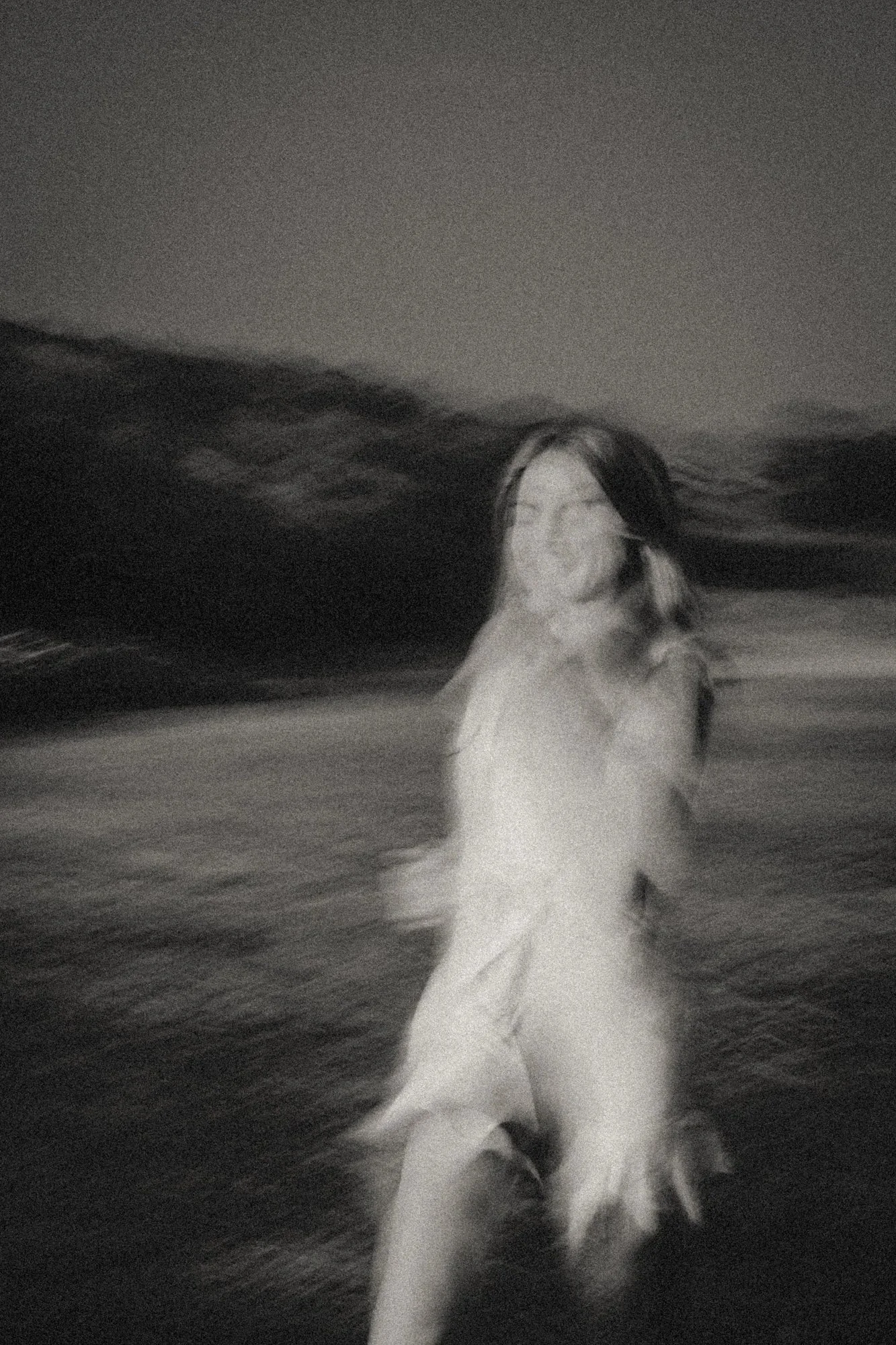 Grainy black-and-white long-exposure photograph of a nude figure moving on a dark shoreline, with wind-blurred hair and a softly misted horizon suggesting dream, tide, and motion.