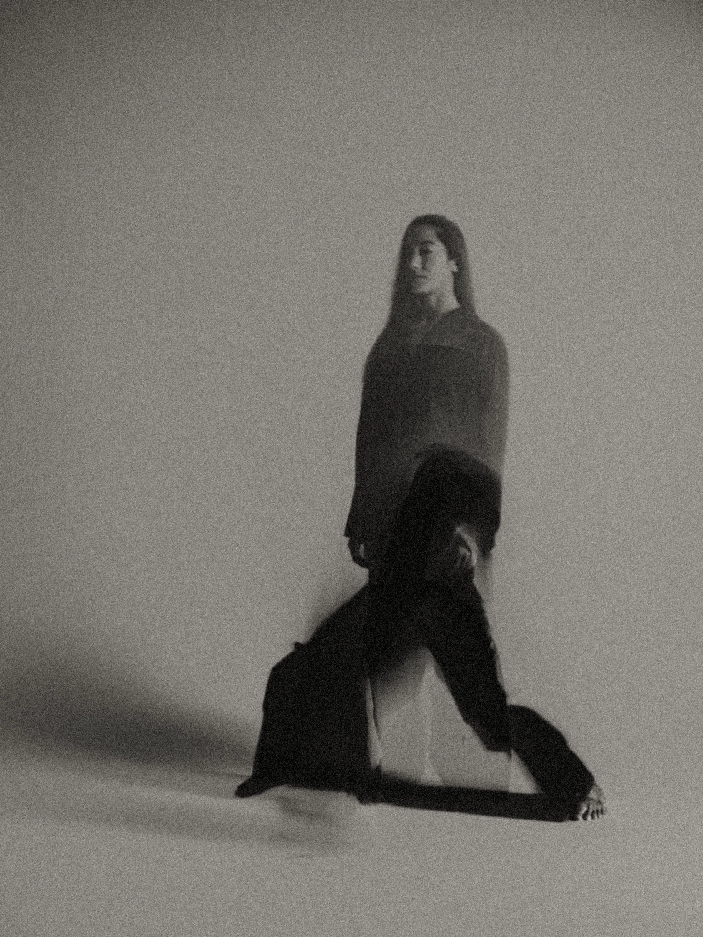 Grainy black-and-white fine art photograph with a double-exposed standing and kneeling figure in a minimalist space, evoking split identity, hierarchy, and the haunting presence of the “double.”