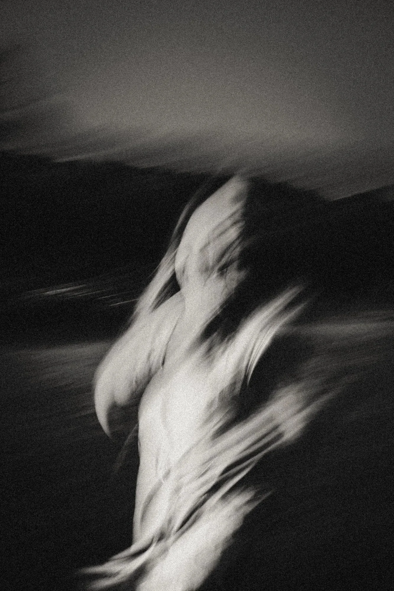 : High-grain black-and-white portrait of a nude figure in heavy motion blur on a dark beach, emphasizing speed, instability, and the body dissolving into night.