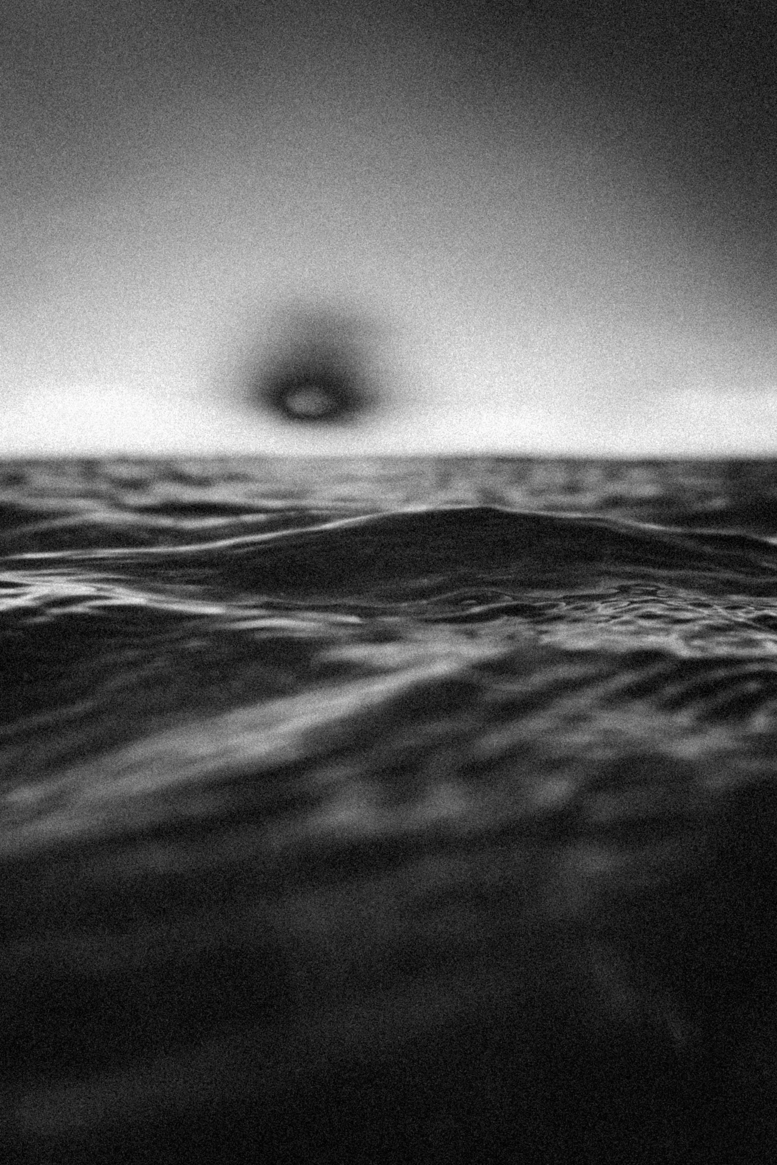 Grainy black-and-white ocean close-up with a low horizon and a dark eclipse-like sun, creating a moody, cosmic seascape.