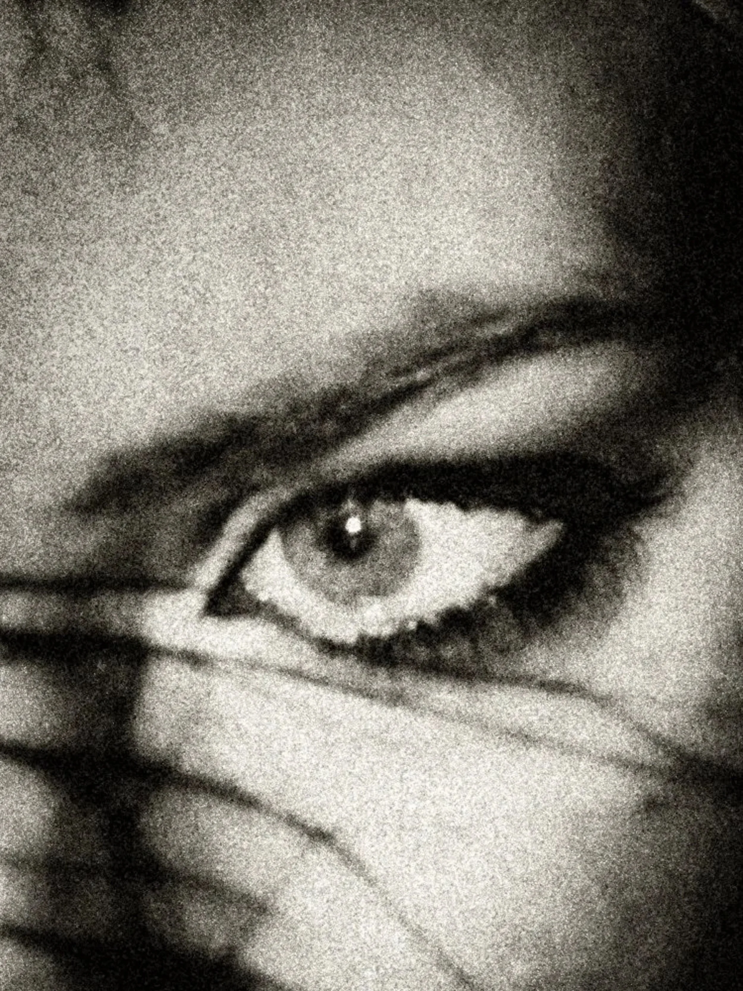 High-grain black-and-white close-up of a single eye partially veiled by shadow or hair, emphasizing intensity, secrecy, and direct gaze.