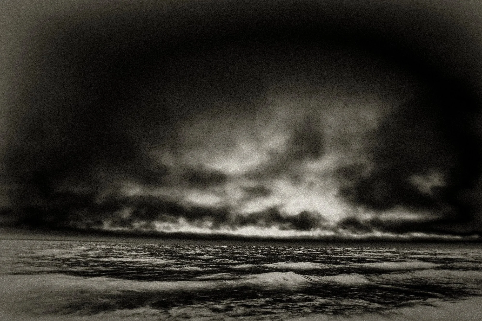 Dramatic inverted black-and-white seascape with heavy storm clouds pressing down on a dim horizon and textured surf in the foreground.