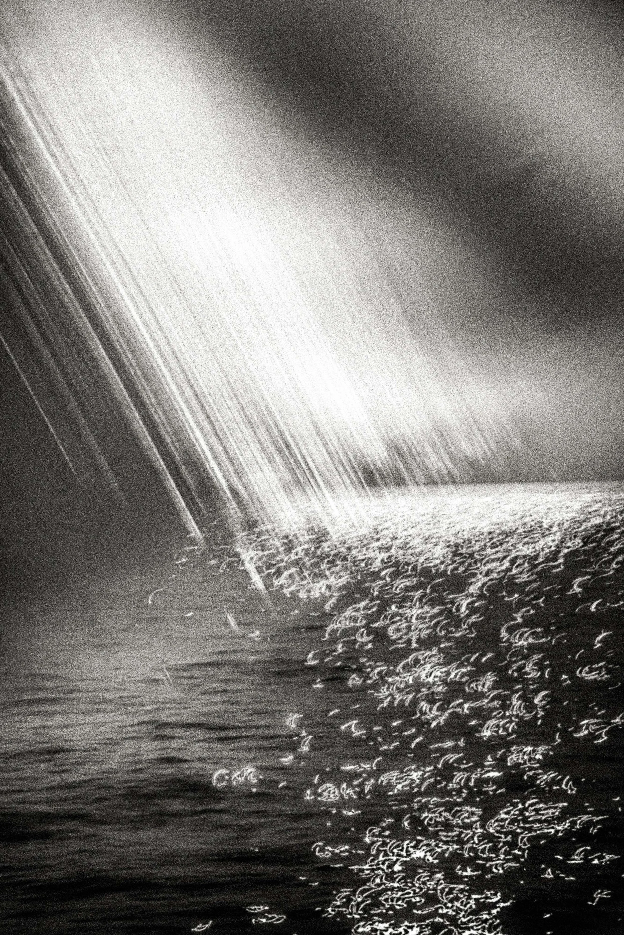 High-grain black-and-white seascape with diagonal rain-like streaks and bright sun-glitter on the water, creating a stormlit field of motion and intensity.