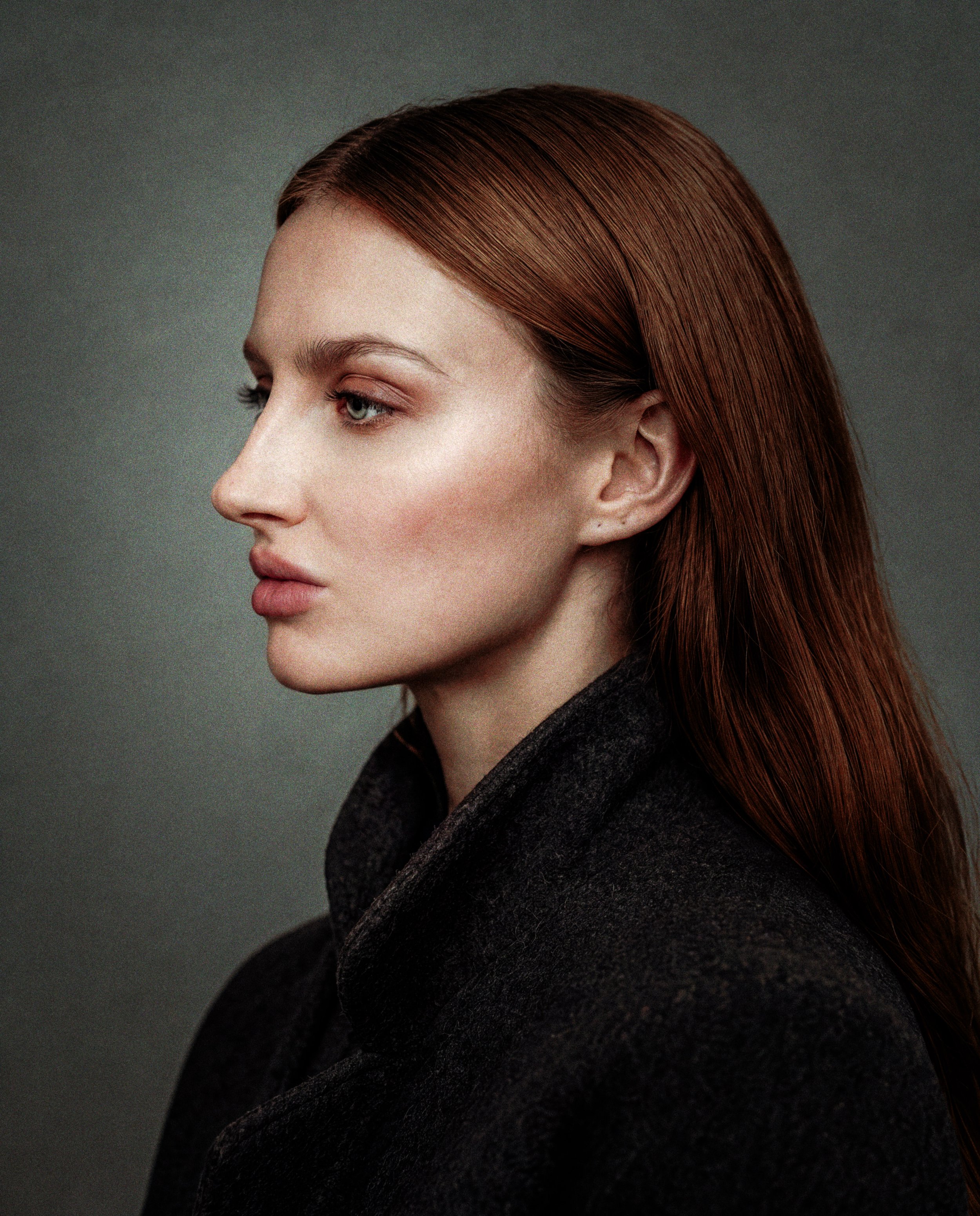 Portrait of a woman with reddish hair, wearing a beige short-sleeve sweater and a black lace skirt, sitting against a dark background with a wooden frame on the right, and wearing a black and amber necklace.
