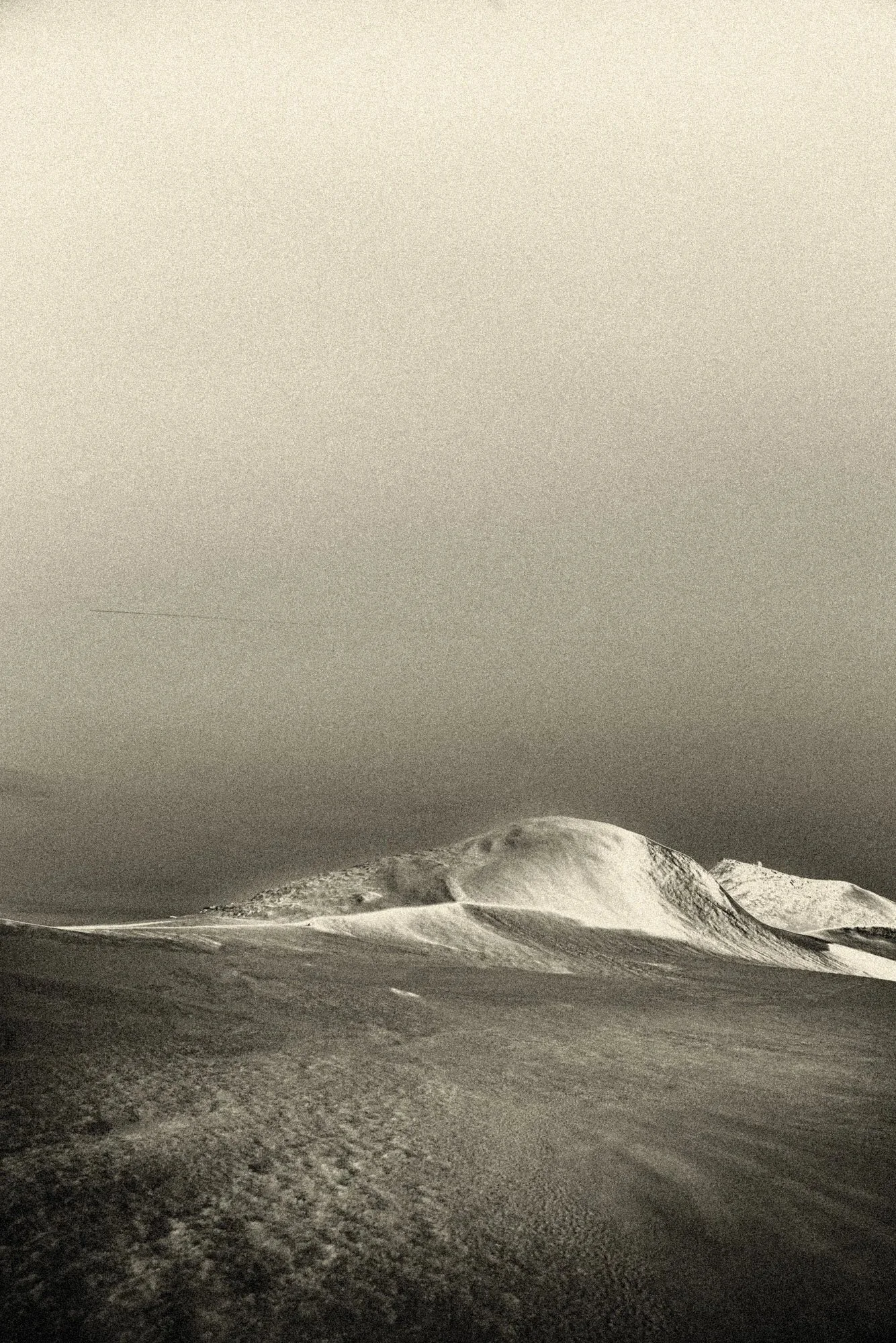 Atmospheric black-and-white snowy ridge line under a vast, pale sky—minimalist fine art landscape photography with soft grain and distant horizons.