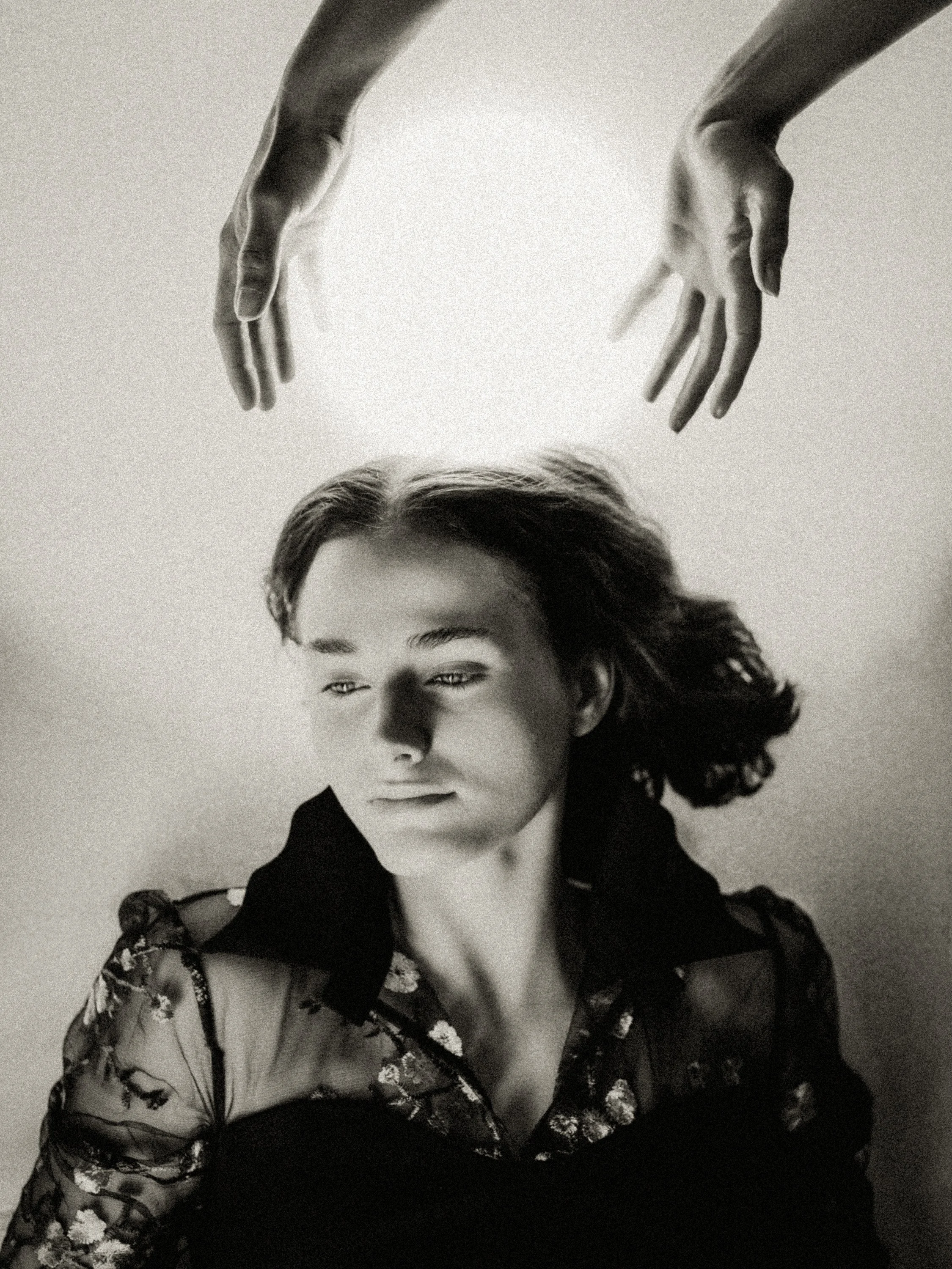Black-and-white portrait of a person lying down while two hands hover above their head, suggesting a blessing or protective gesture.