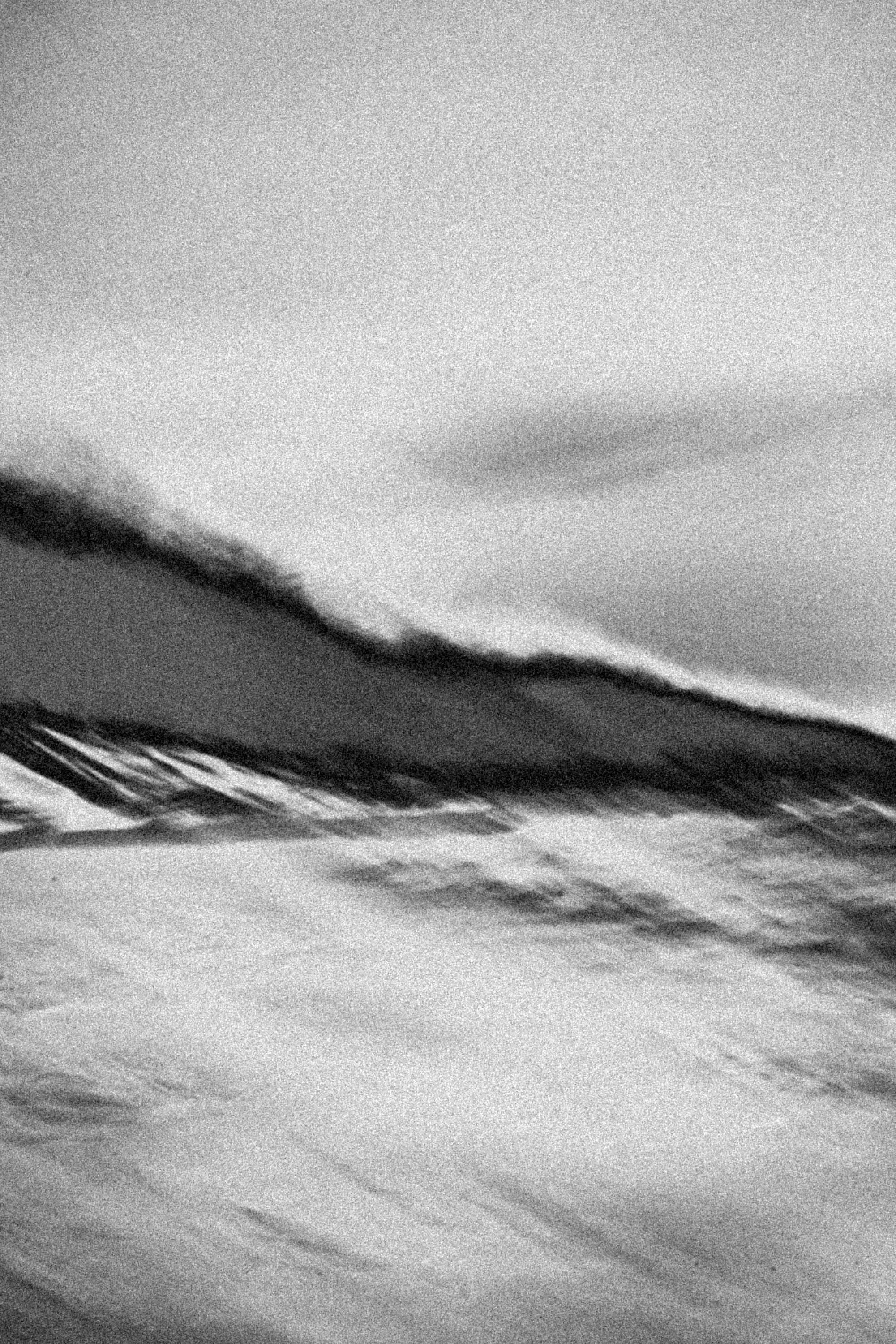 Grainy monochrome seascape abstraction with a blurred dark ridge and wind-swept horizon, capturing motion, salt air, and the sea’s slow, spectral pull.