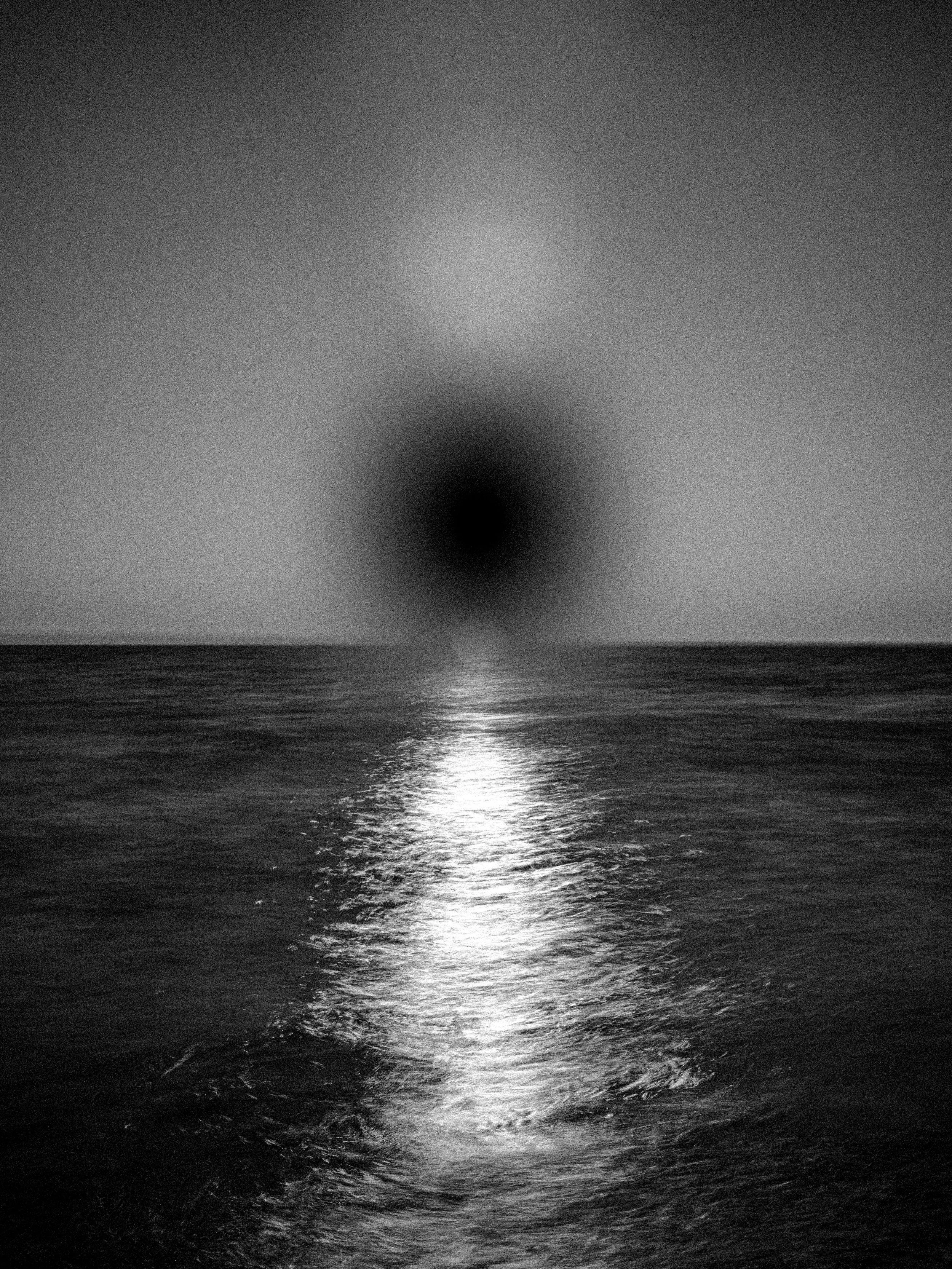 Grainy black-and-white seascape with a dark eclipse-like disc over the horizon and a bright vertical reflection path on rippling water, conveying cosmic solitude and nocturnal atmosphere.