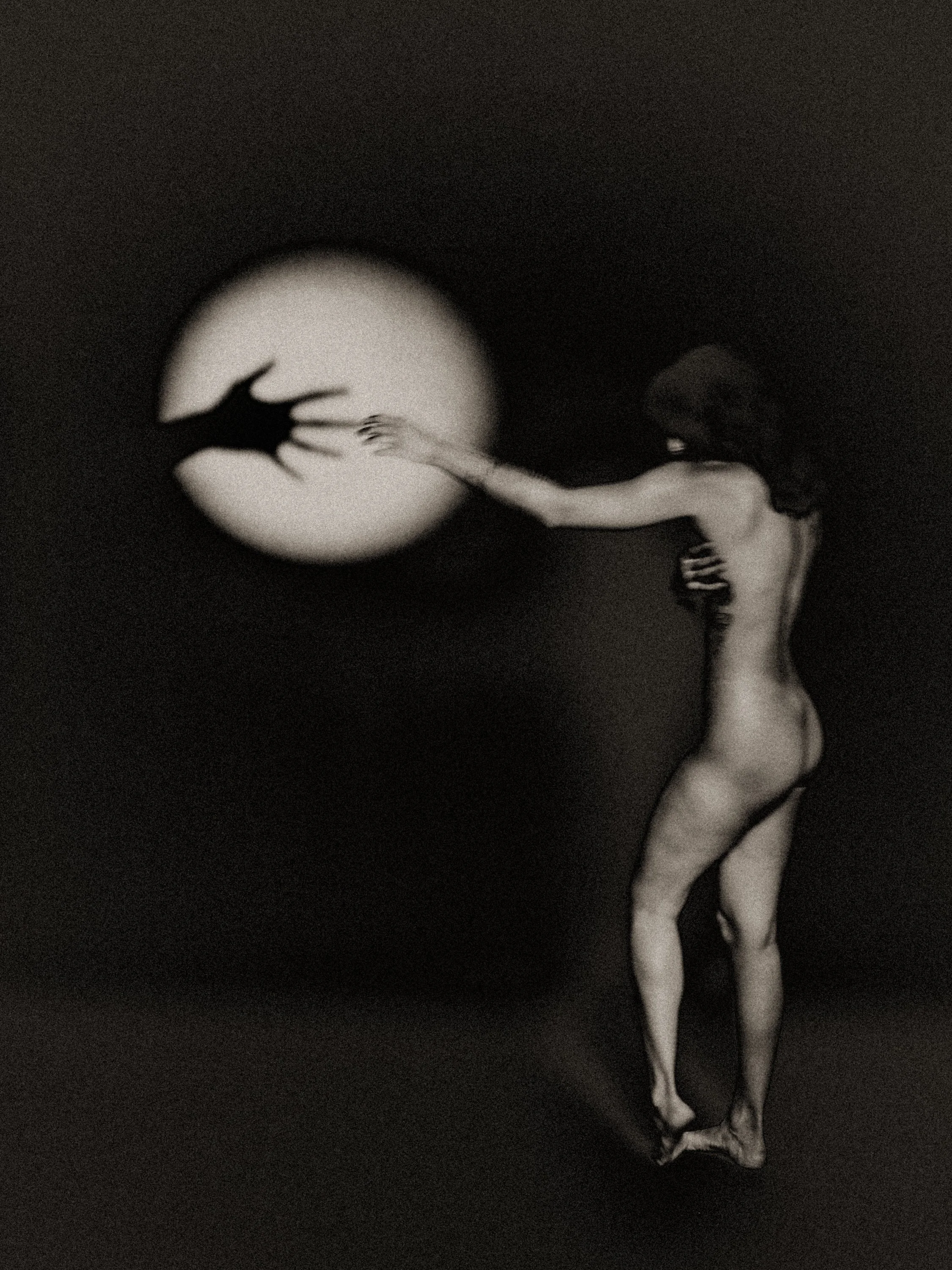 Surreal black-and-white nude figure reaching toward a glowing moon-like disc with a shadowed hand inside—fine art photography about longing, touch, and the distance between bodies and light.