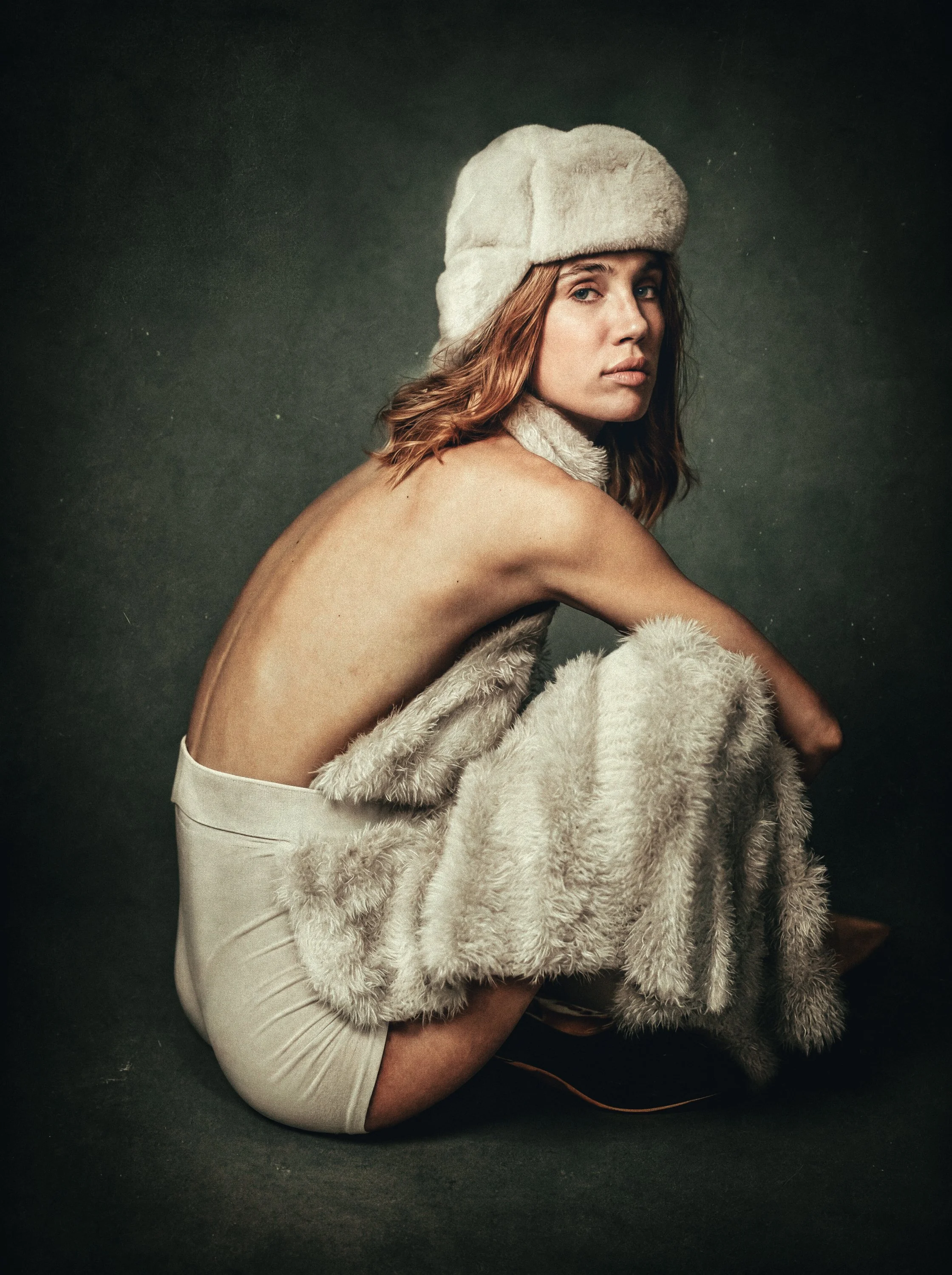 Black and white photo of a woman sitting in a curled position, wearing a large fur hat, fur gloves, and high-heeled shoes, with her back partially exposed.