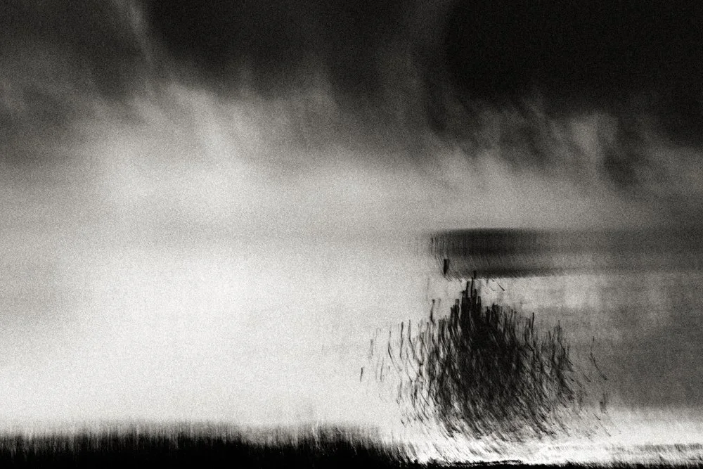 Stormy black and white seascape with swirling fog and a dark clustered form near the horizon, dramatic atmospheric coastal abstraction.