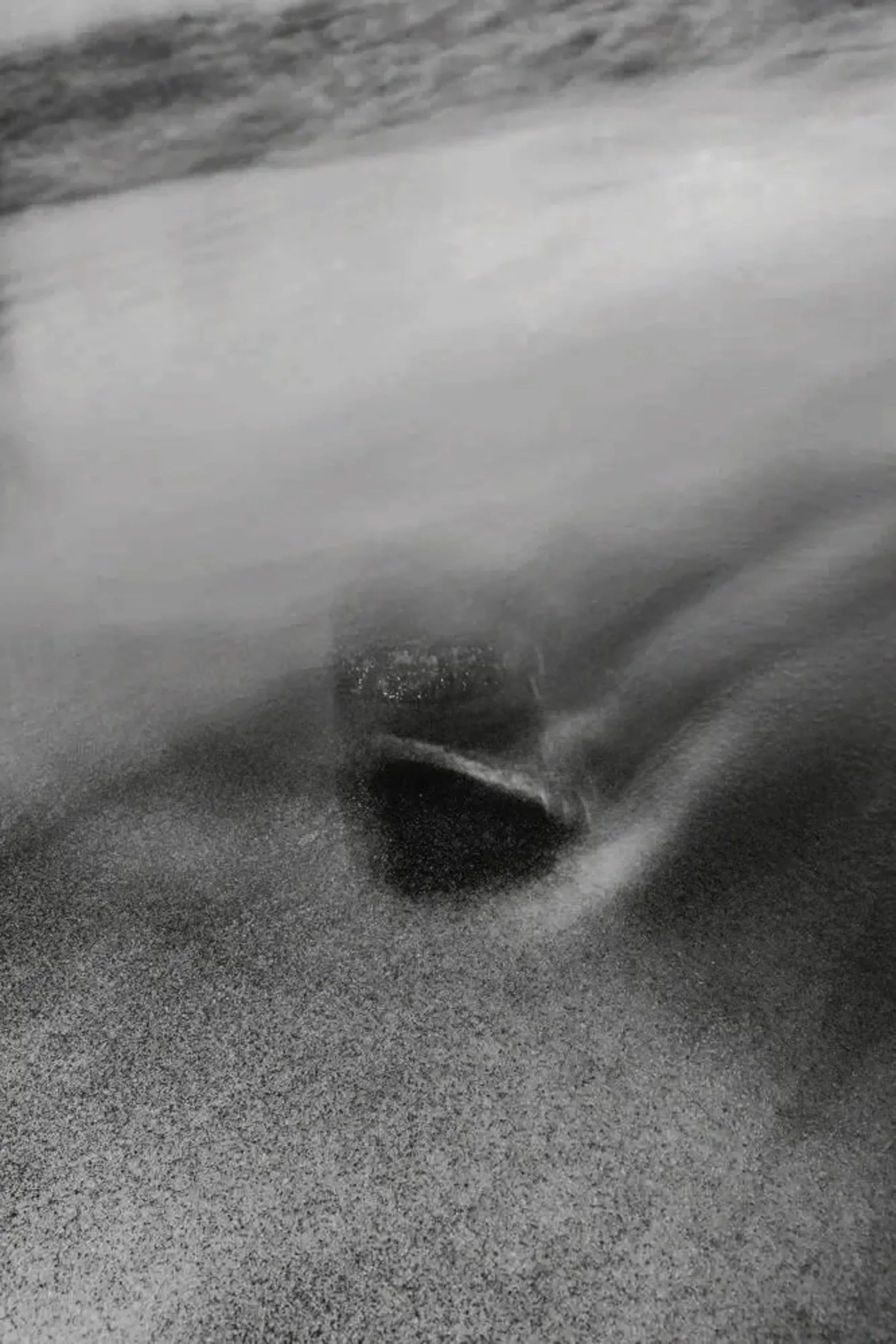 Abstract monochrome shoreline where water sweeps around a small dark rock set into wet sand, suggesting erosion, disappearance, and the pull of tide.