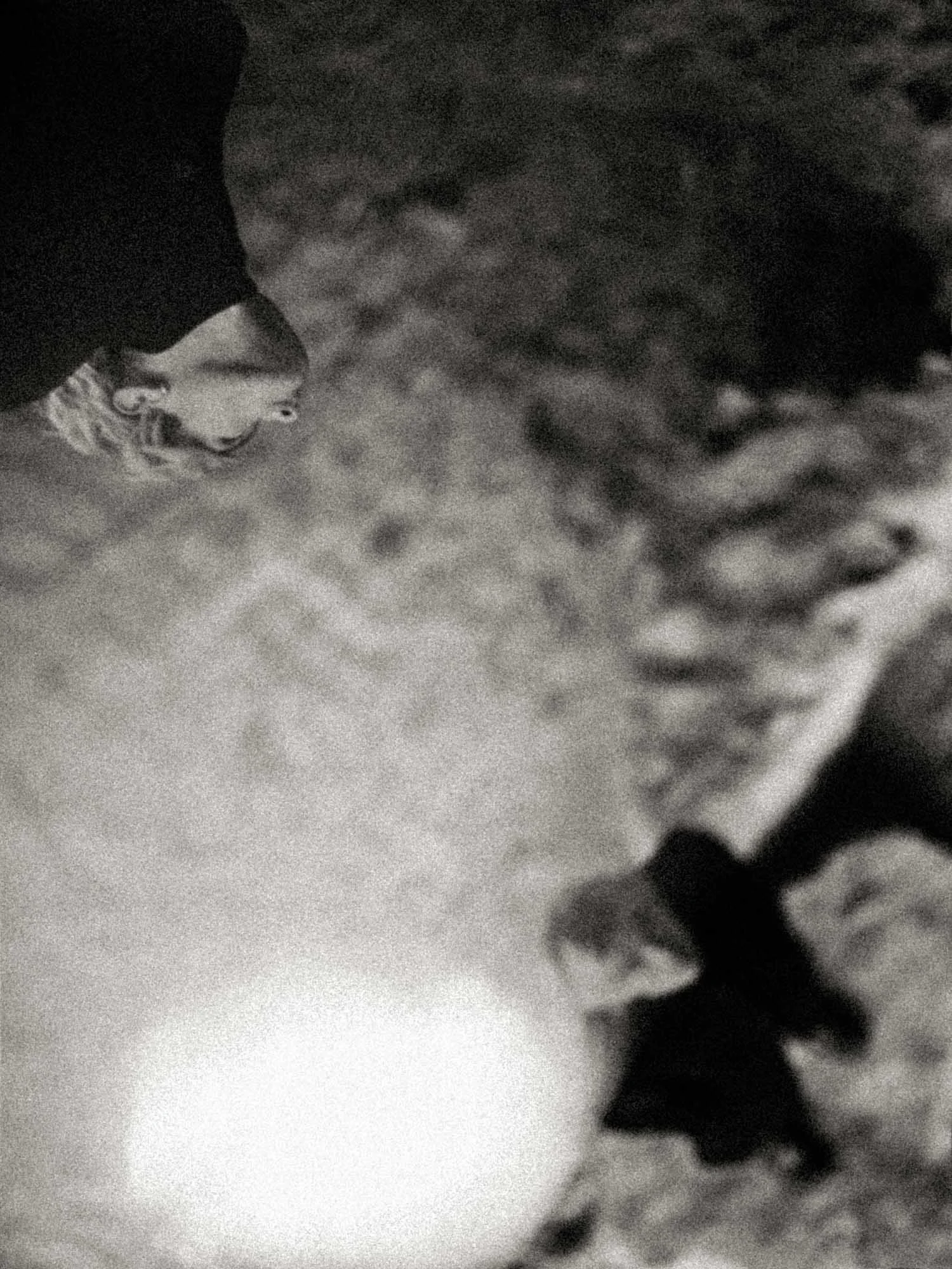 Grainy black-and-white fine art photograph with an inverted, drifting face and a glowing circular light source, evoking sleep, disorientation, and dream-illumination.