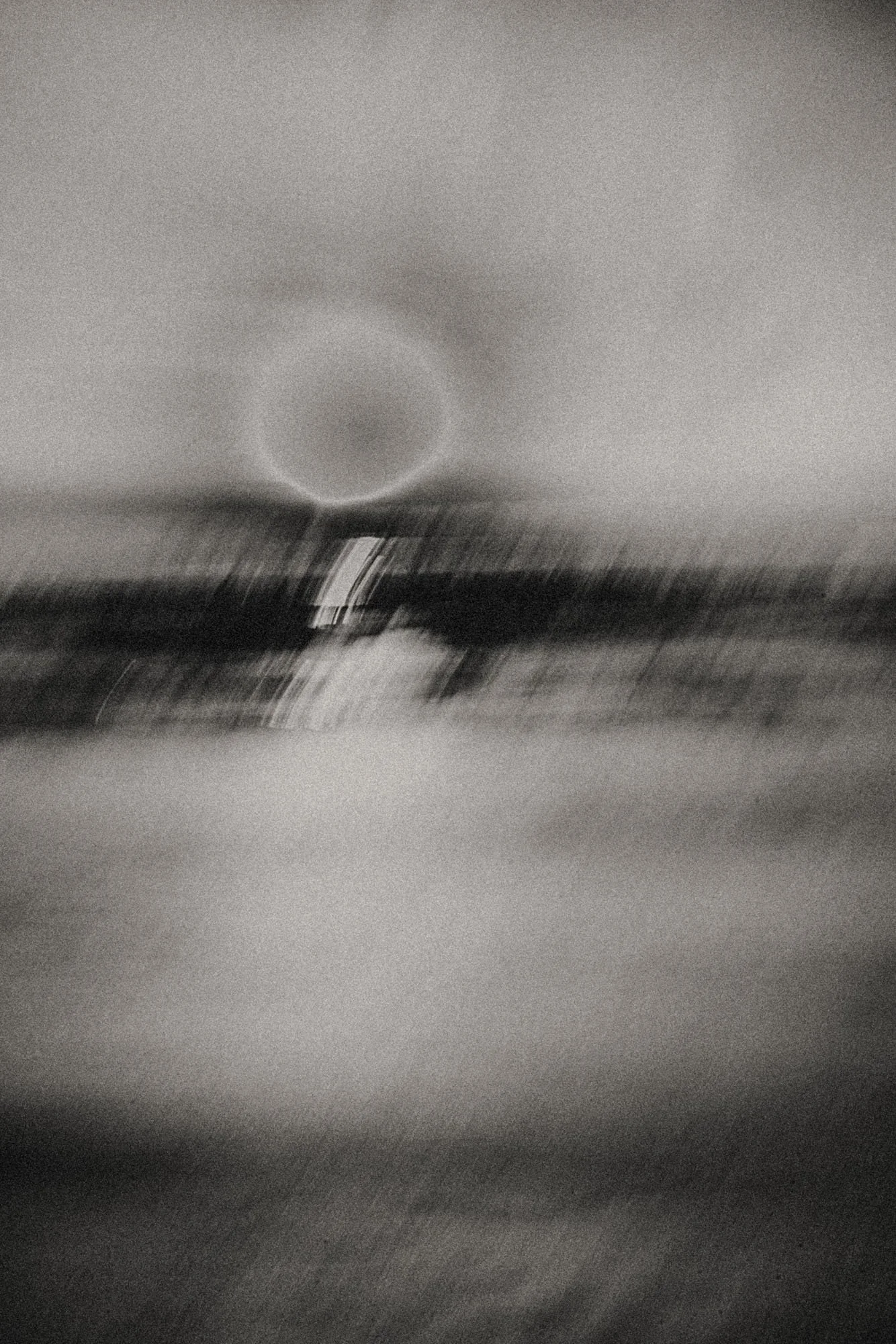 Grainy black-and-white fine art seascape with a soft eclipsed circle above the horizon and a luminous vertical reflection, suggesting solitude, vastness, and night-lit water.