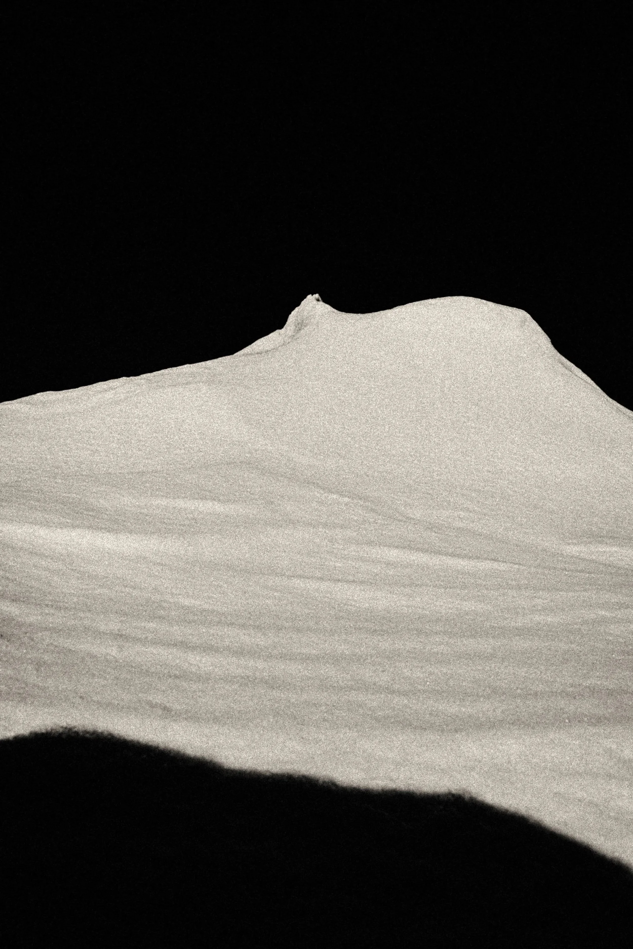 Minimalist, high-grain black-and-white fine art photograph of a snow ridge or dune against a deep black sky, emphasizing texture, quiet, and negative space.