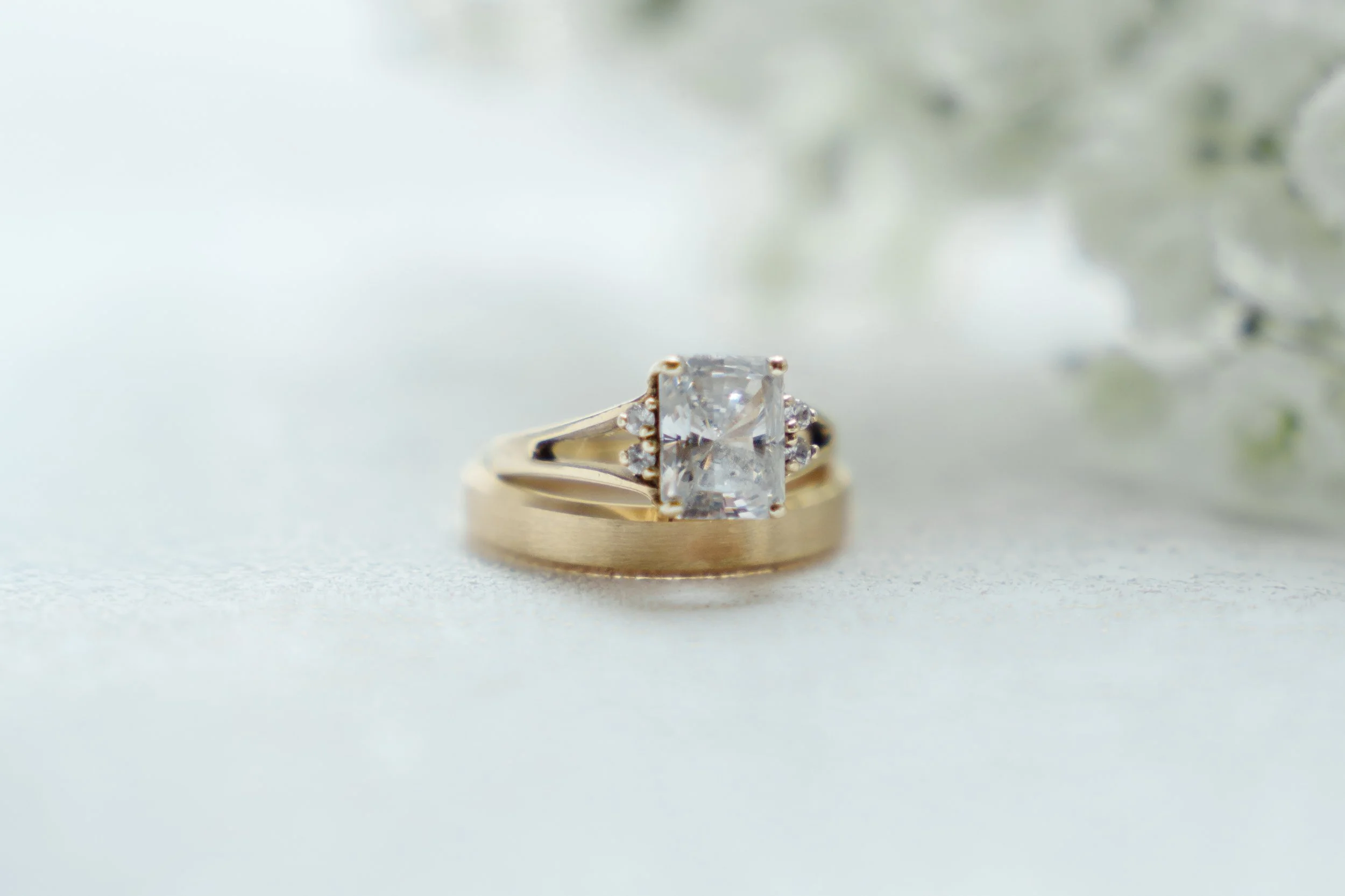 Close-up of a gold engagement ring with a rectangular diamond center stone and small side diamonds, placed on a white textured surface with soft focus background.