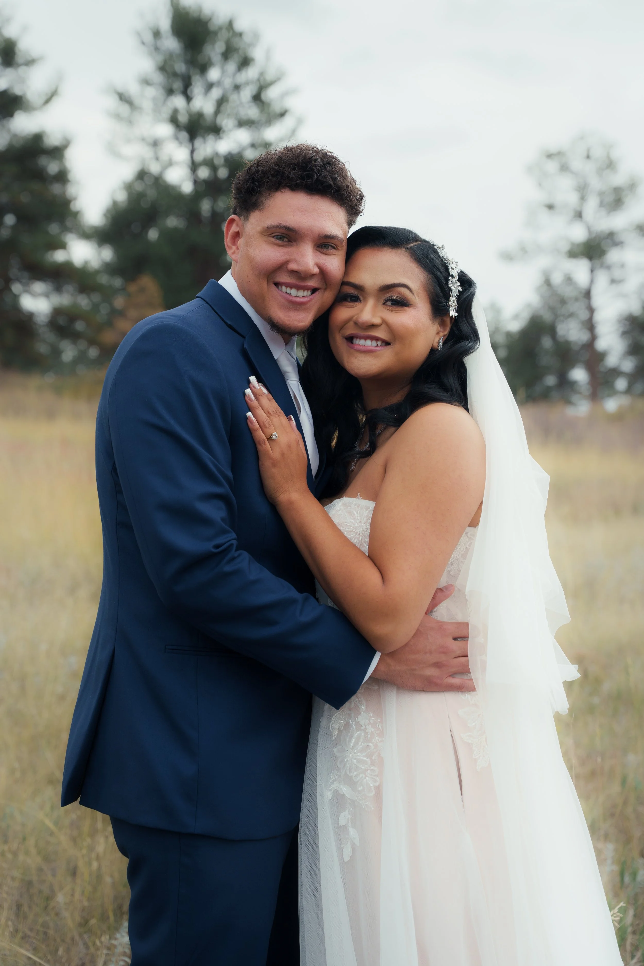 A smiling couple, dressed in wedding attire, embracing outdoors in a grassy field with trees in the background.