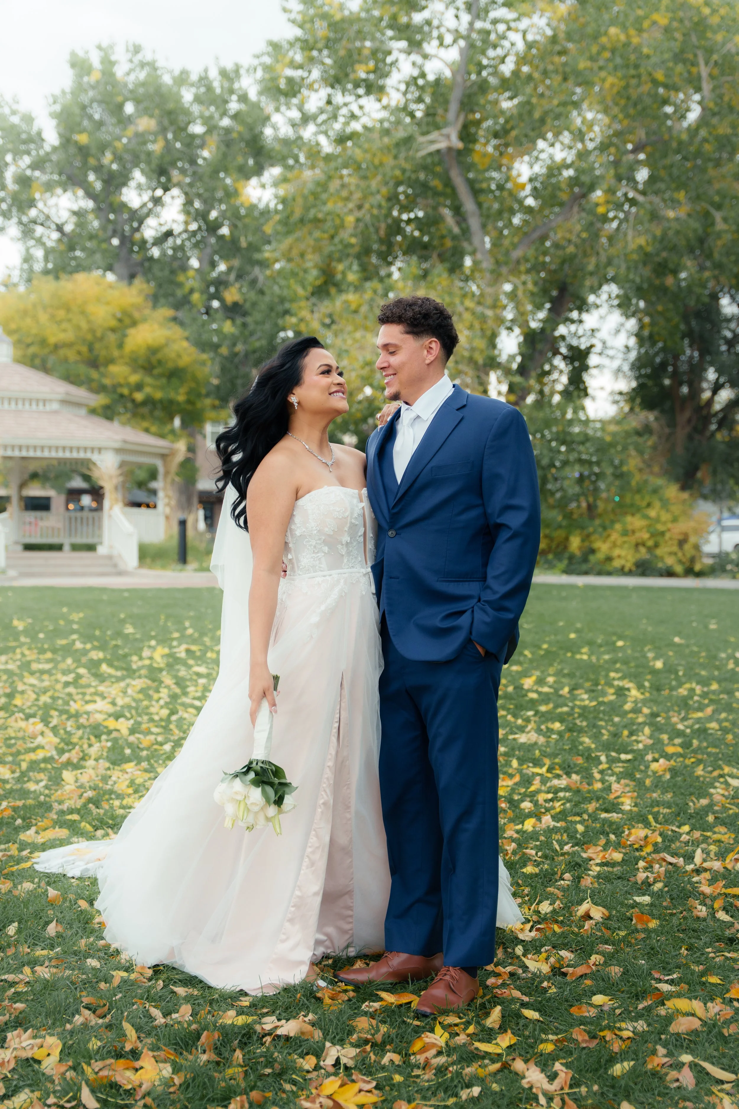 A bride and groom smiling at each other outdoors on a grassy field with fallen yellow leaves, trees, and a gazebo in the background.