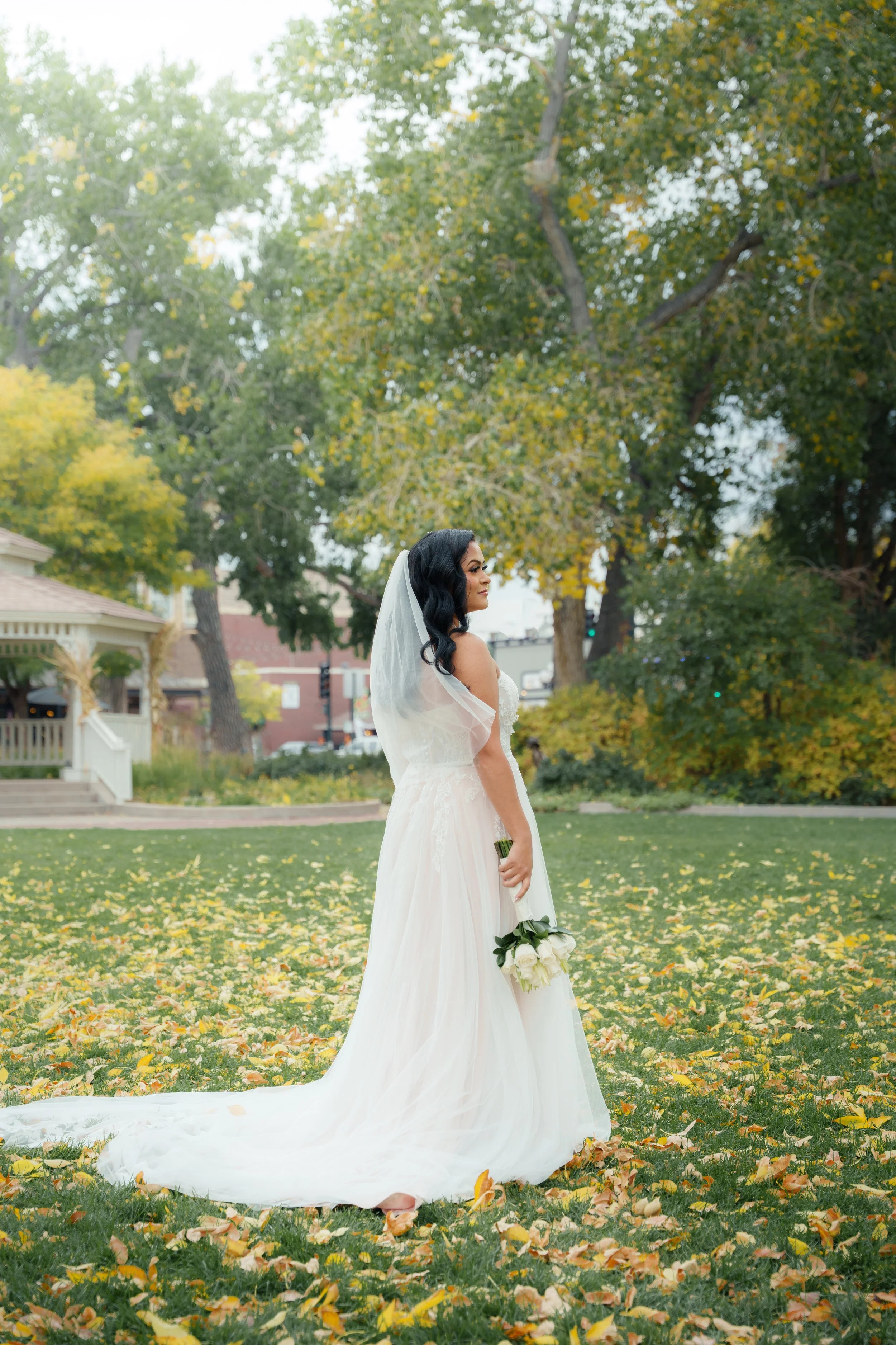 A bride in a white wedding dress with black hair and a veil holding a bouquet of white roses in a park with trees and fallen leaves.